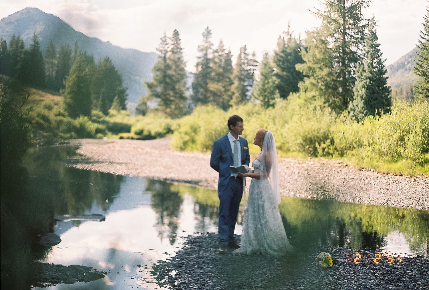 Bride and groom exchanging vows beside a serene mountain stream surrounded by pine trees.