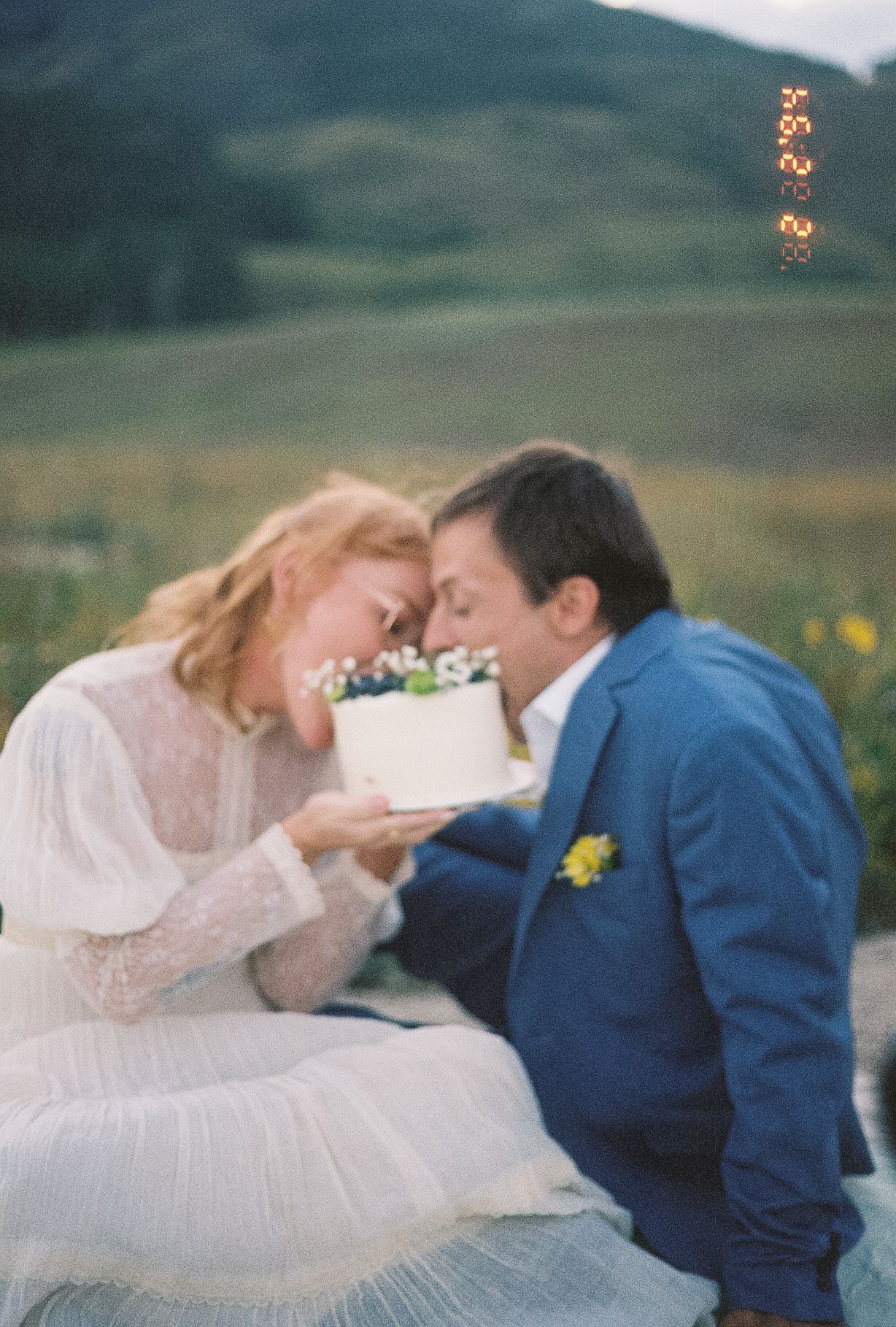 Bride and groom touch foreheads while sharing a small white wedding cake outdoors in a mountain setting.