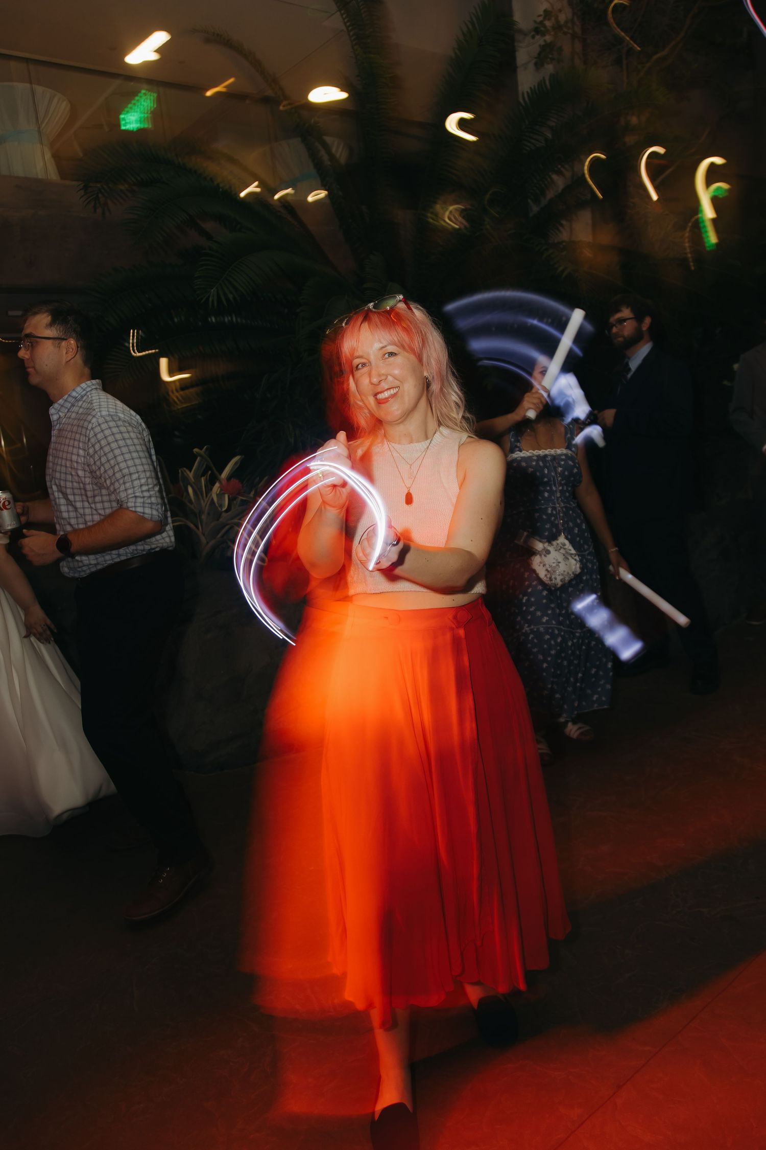Guest in orange skirt dances with glowing light wands at vibrant wedding reception party.