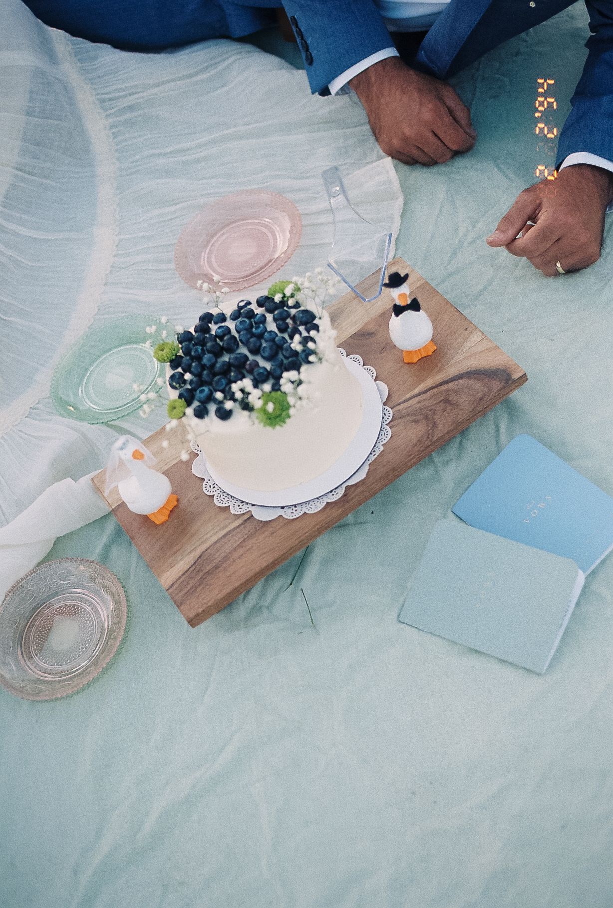 Elegant wedding cake topped with fresh blueberries on a wooden board, surrounded by pastel glassware on a light blue cloth.