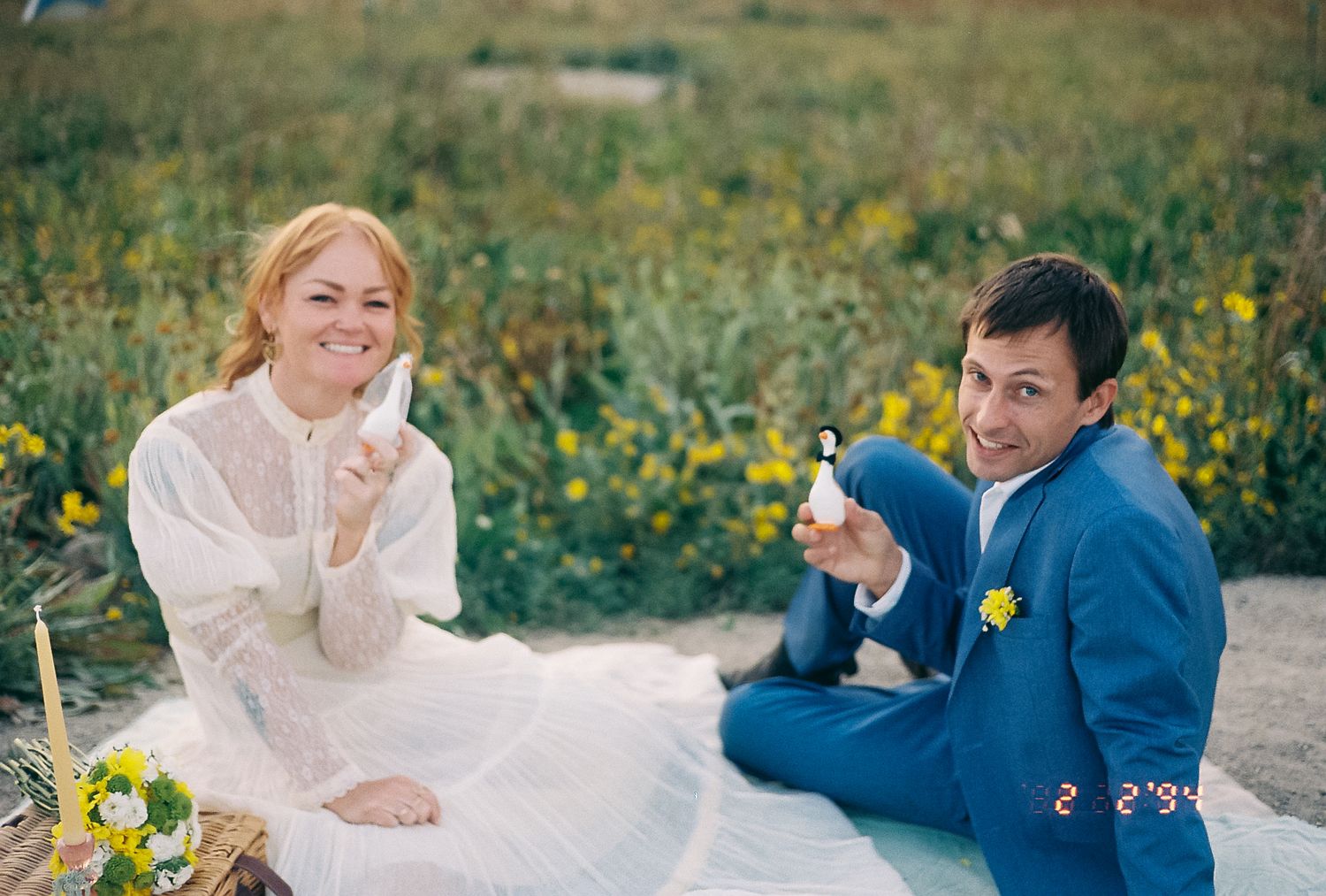 A smiling bride in white dress and groom in blue suit sit in a flower field holding small wedding figurines.