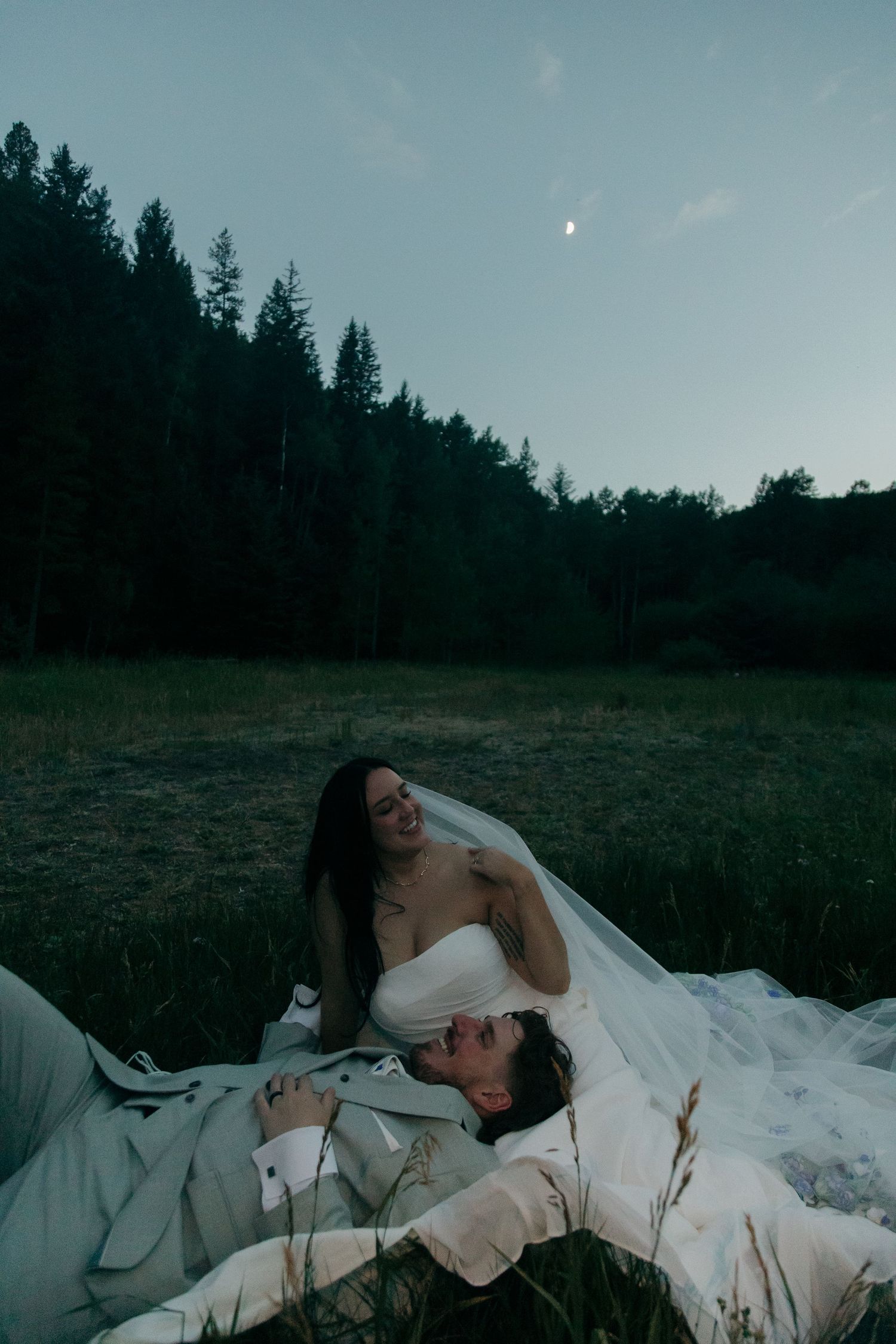 Bride smiles at groom lying in her veil in a twilight meadow with crescent moon overhead.