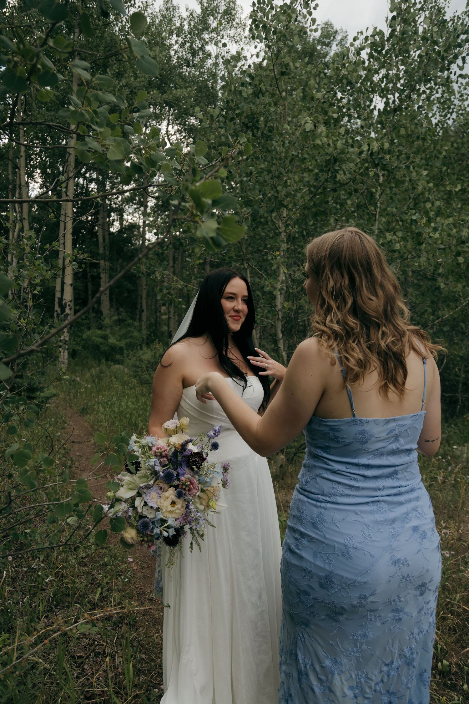 Bride in white gown holds wildflower bouquet while bridesmaid in blue dress adjusts her dress in a forest setting.