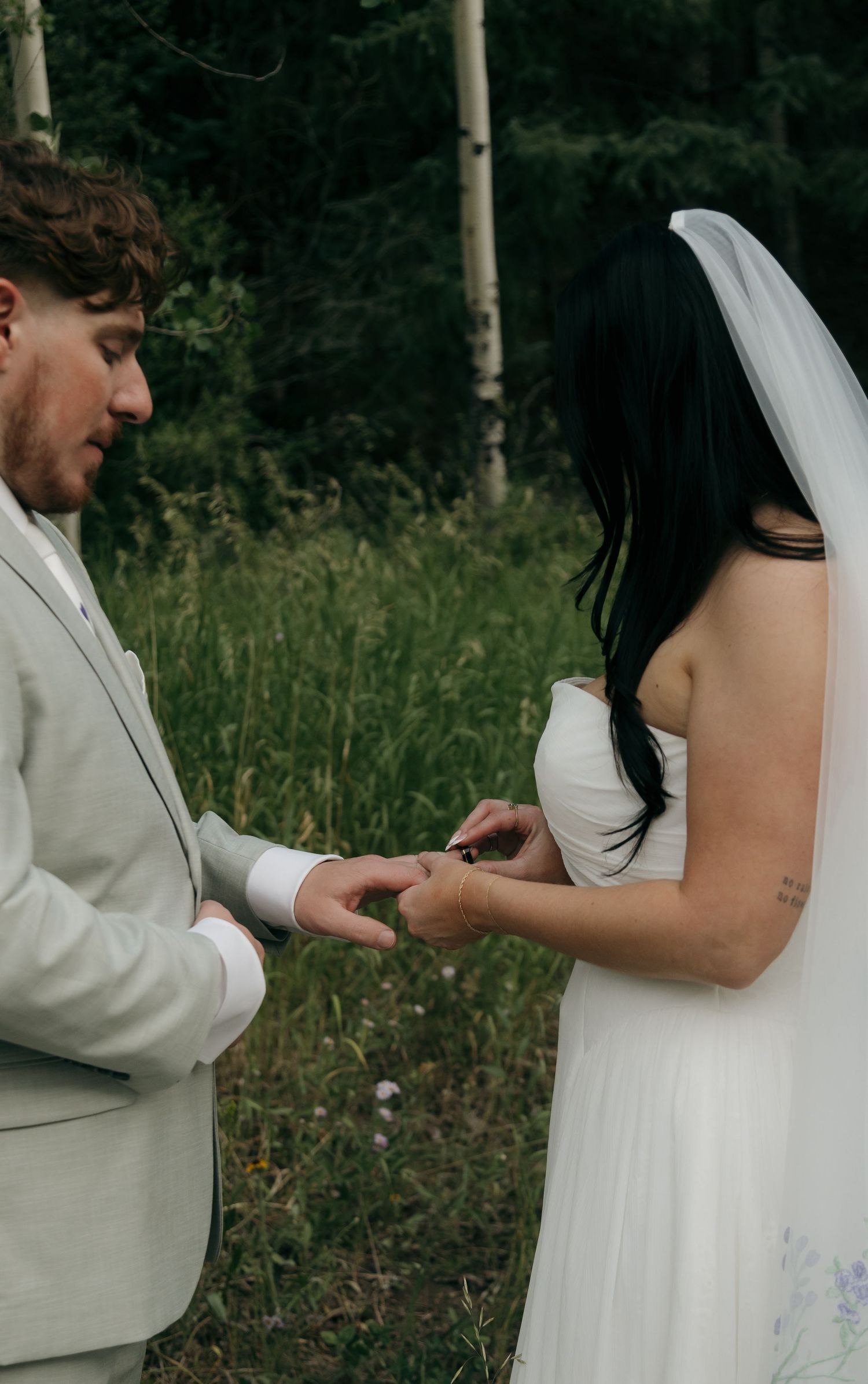 Groom places wedding ring on bride's finger during intimate outdoor forest elopement ceremony.