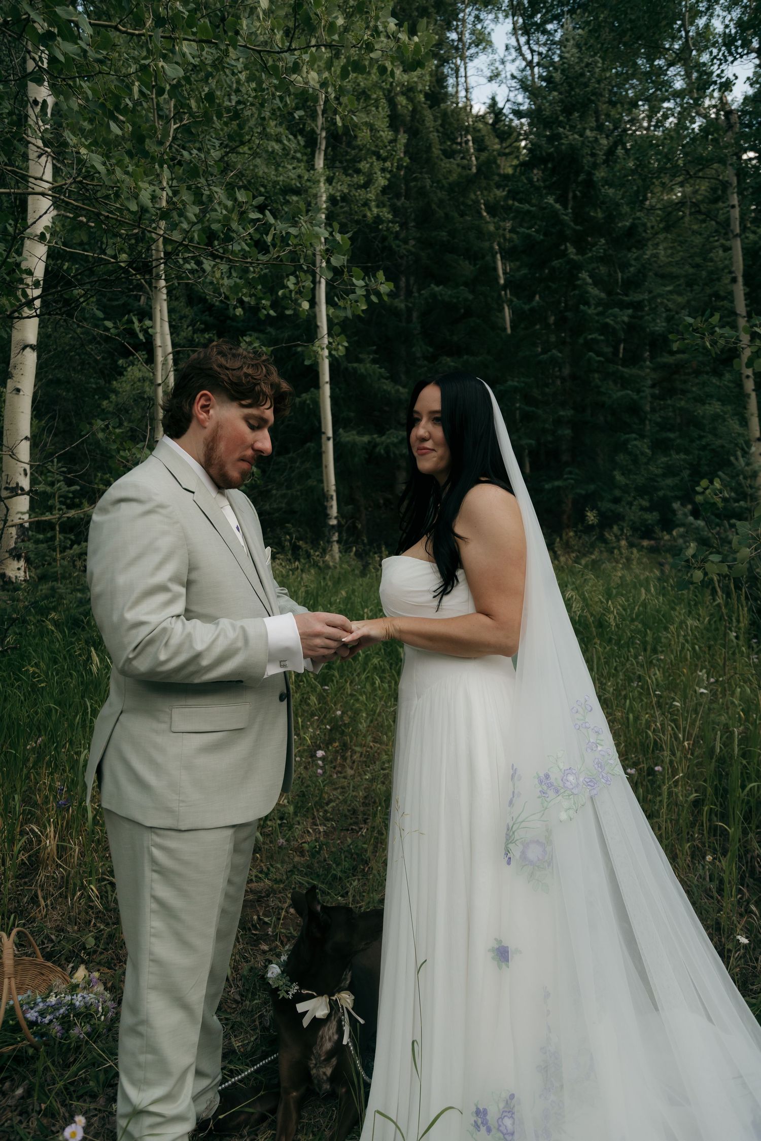 Groom places ring on bride's finger during intimate outdoor forest elopement ceremony with dog nearby.