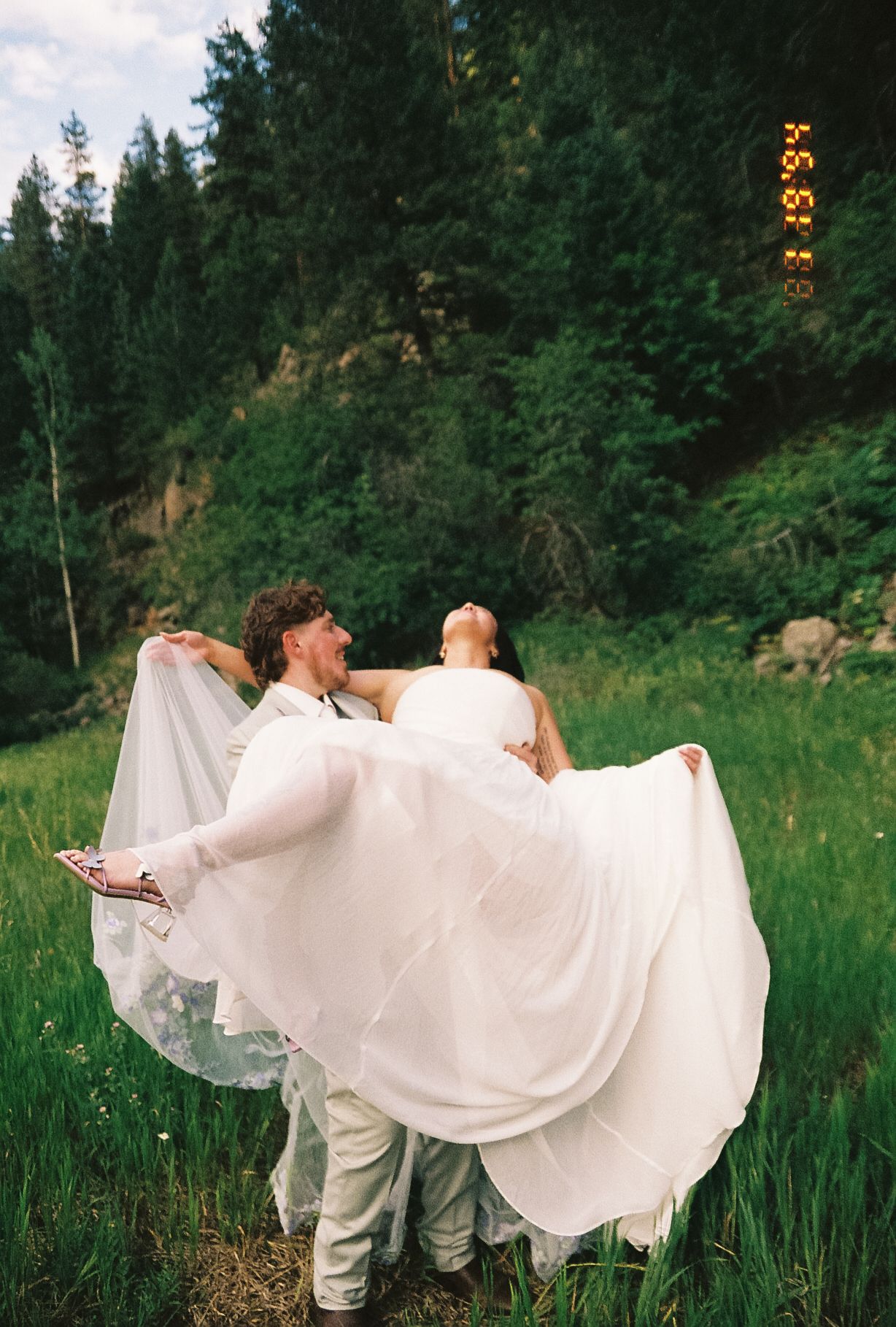 Groom lifts and twirls bride in flowing white veil and gown in a lush green forest clearing.