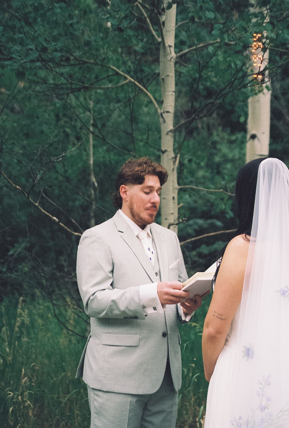 Groom in grey suit reading wedding vows to bride in forest elopement ceremony on film.