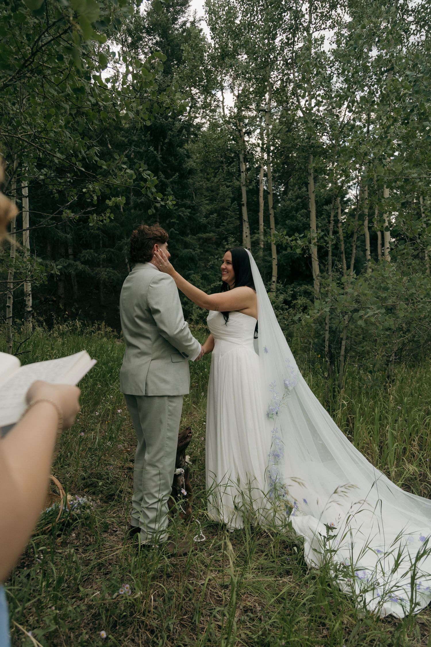 Bride tenderly touches grooms face during romantic outdoor forest wedding ceremony with flowing floral cathedral veil.