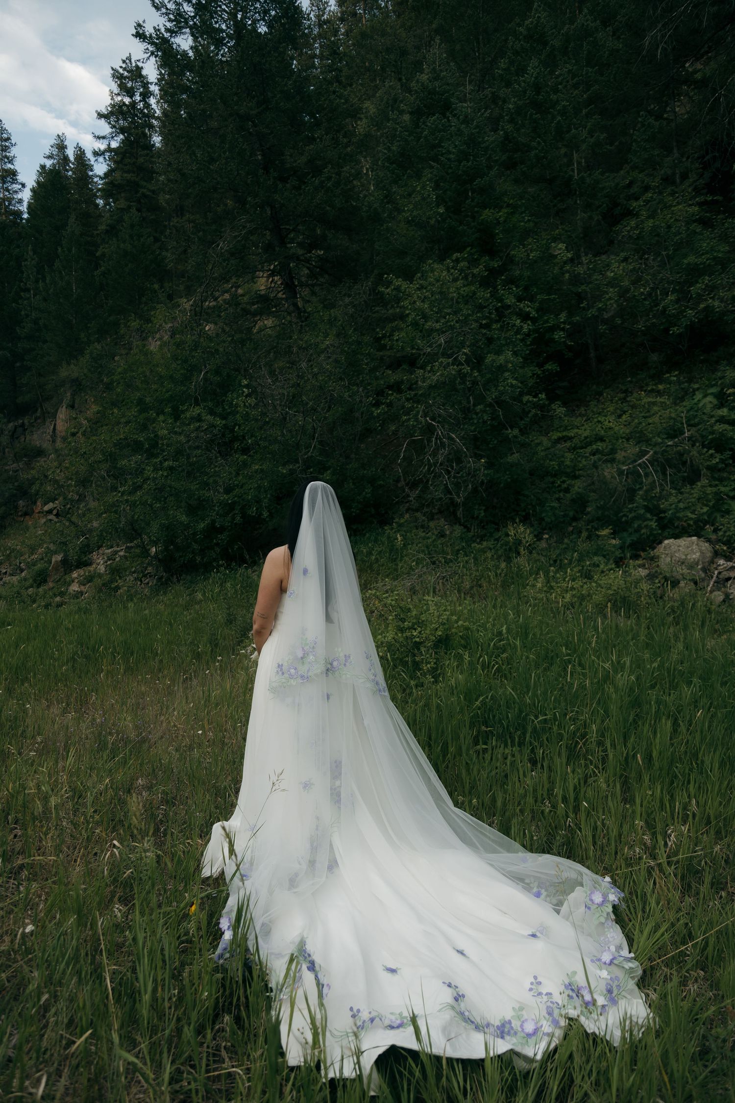 Bride in white gown with floral embroidered veil trails through lush green mountain meadow.