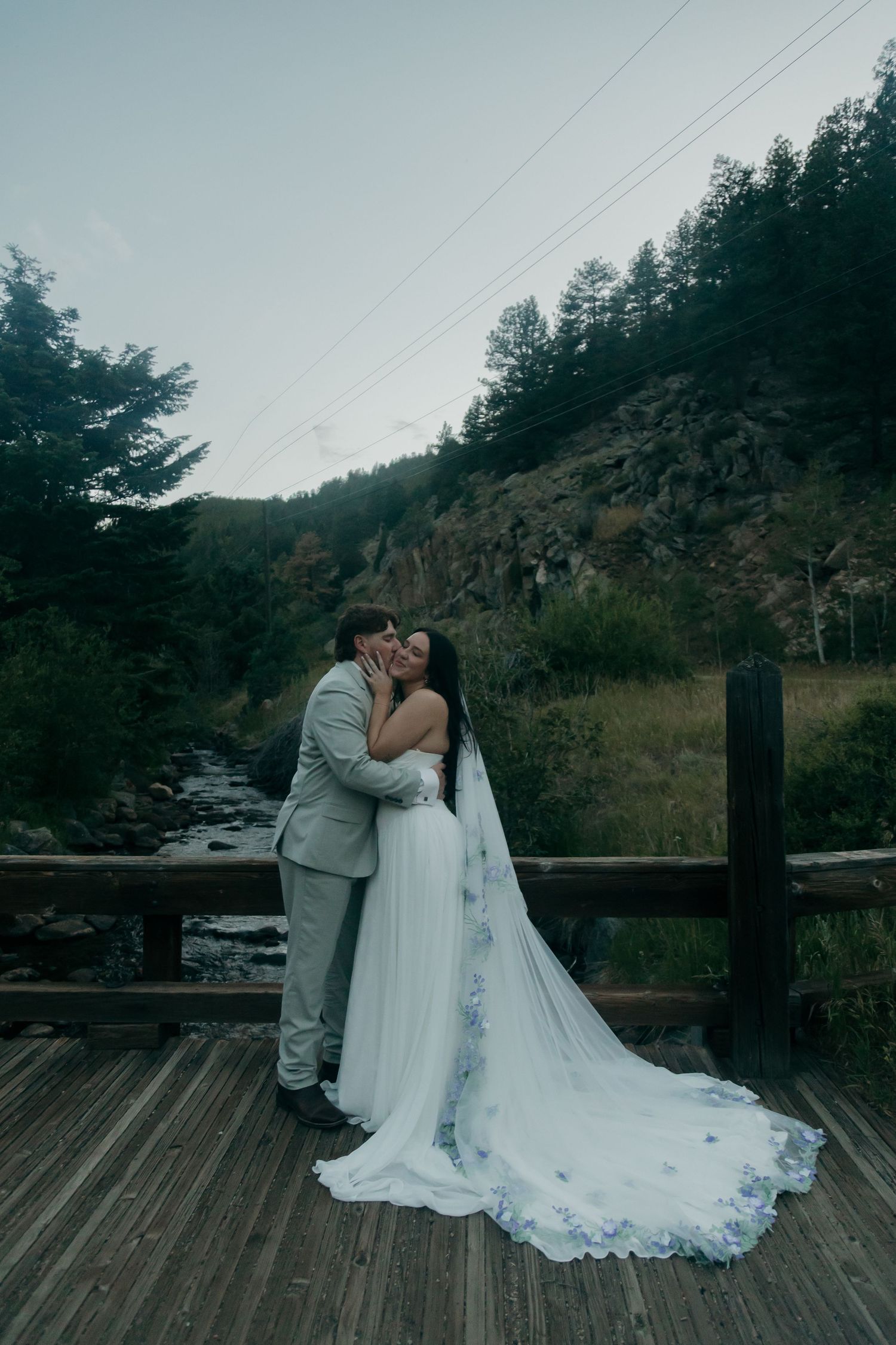Bride and groom share a kiss on a wooden bridge with rocky mountain stream and forest behind.
