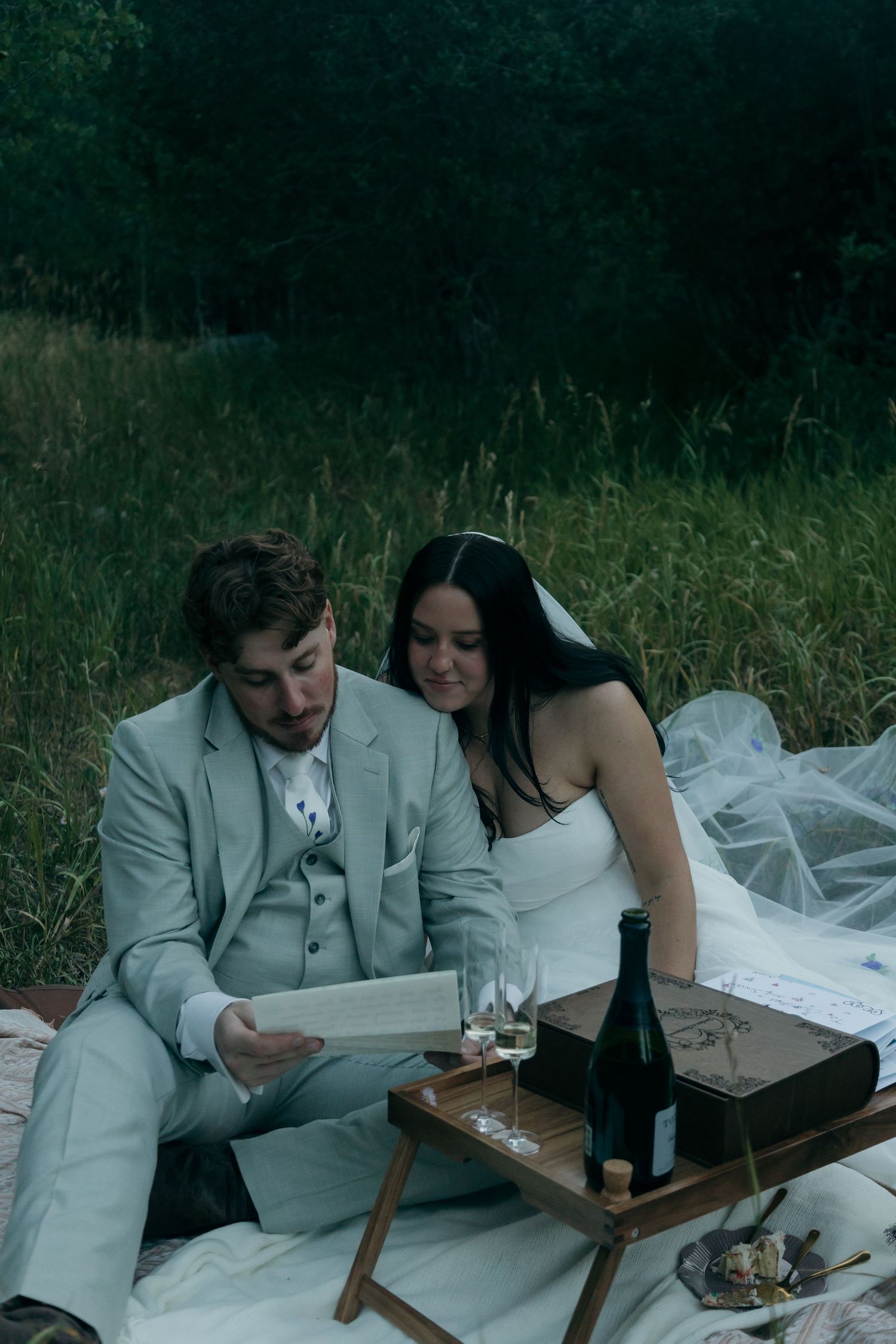 Newlyweds reading together at an outdoor wedding picnic with champagne on a wooden tray.