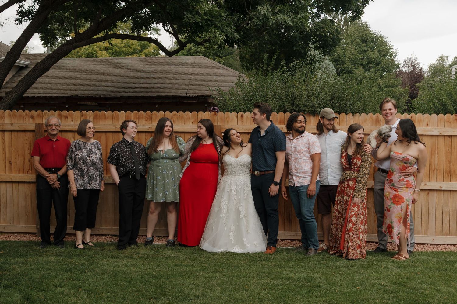 Outdoor wedding reception guests gather in backyard setting with wooden fence and trees in background.