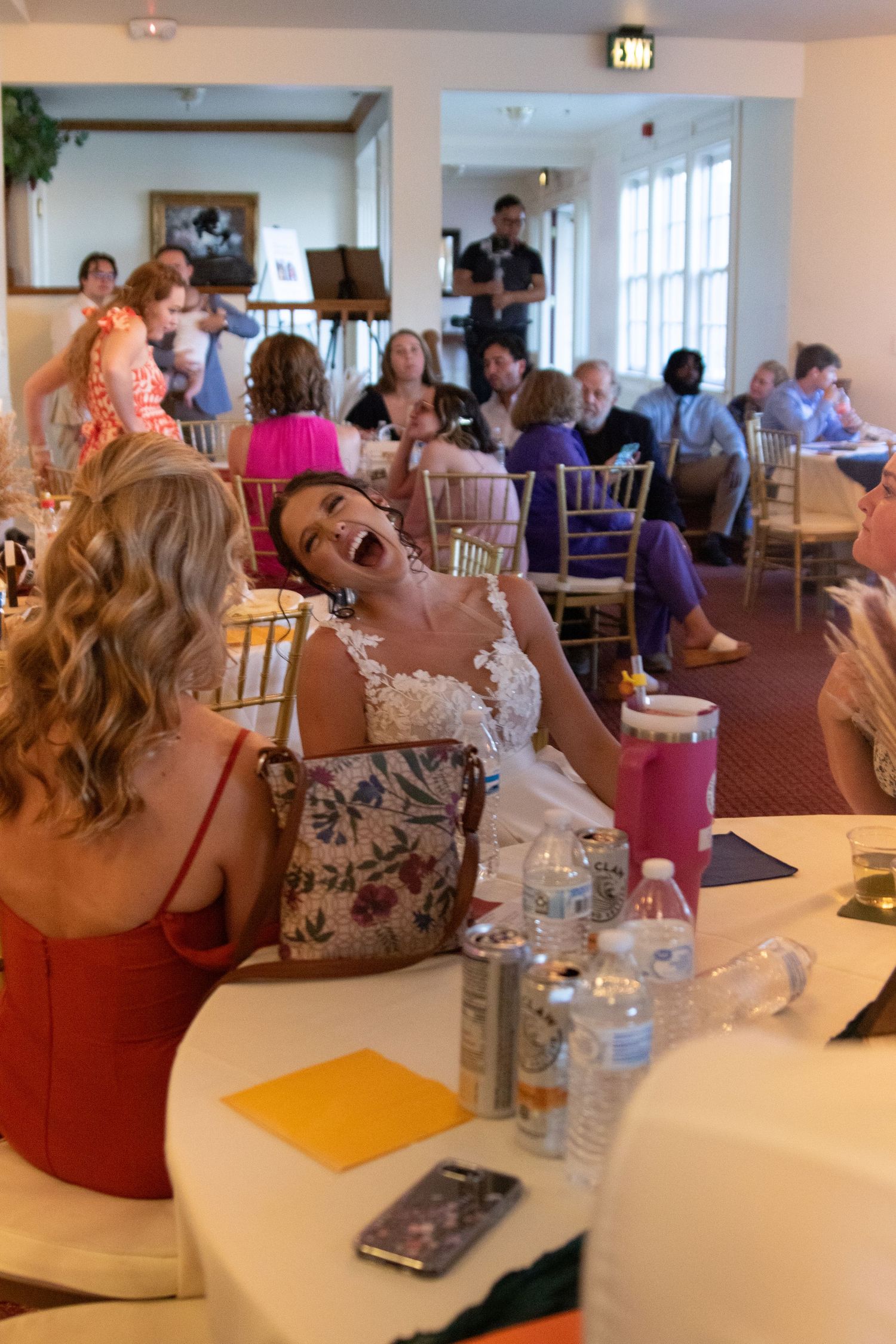 Wedding reception guests laughing and enjoying themselves at tables in a bright venue.