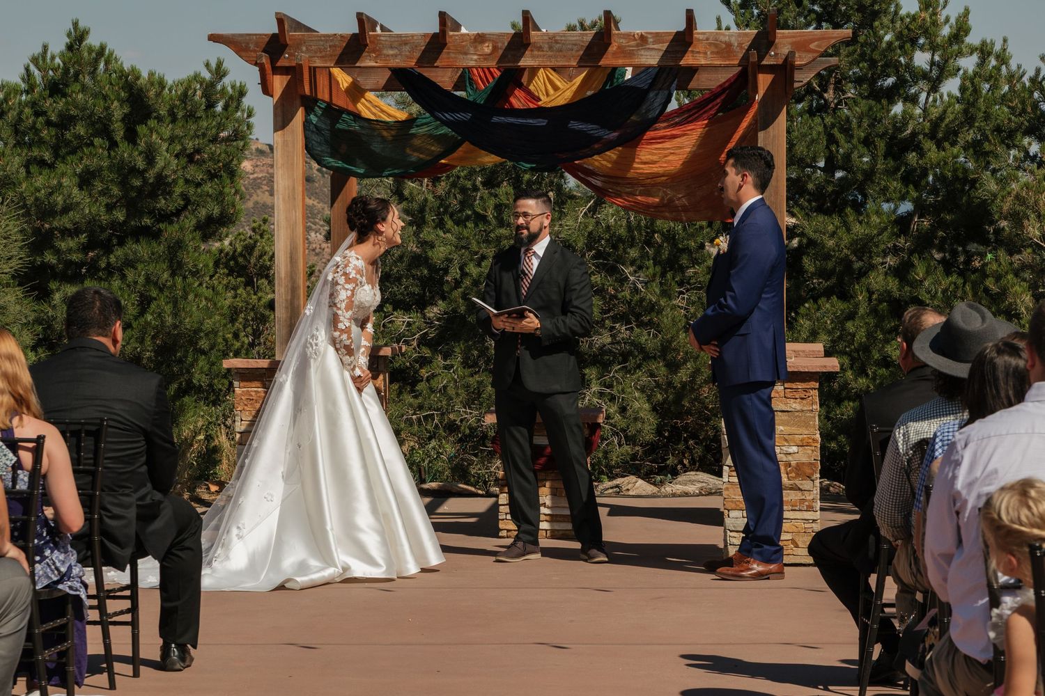 Outdoor wedding ceremony under a wooden pergola decorated with fabric draping.