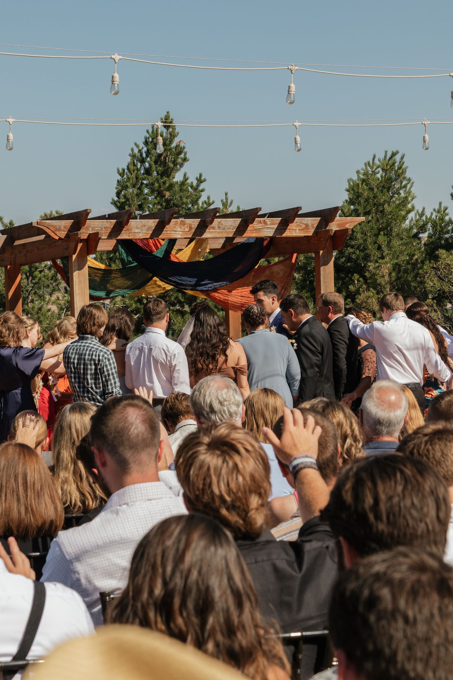 Wedding guests gather outdoors under string lights and a wooden pergola during a sunny ceremony.