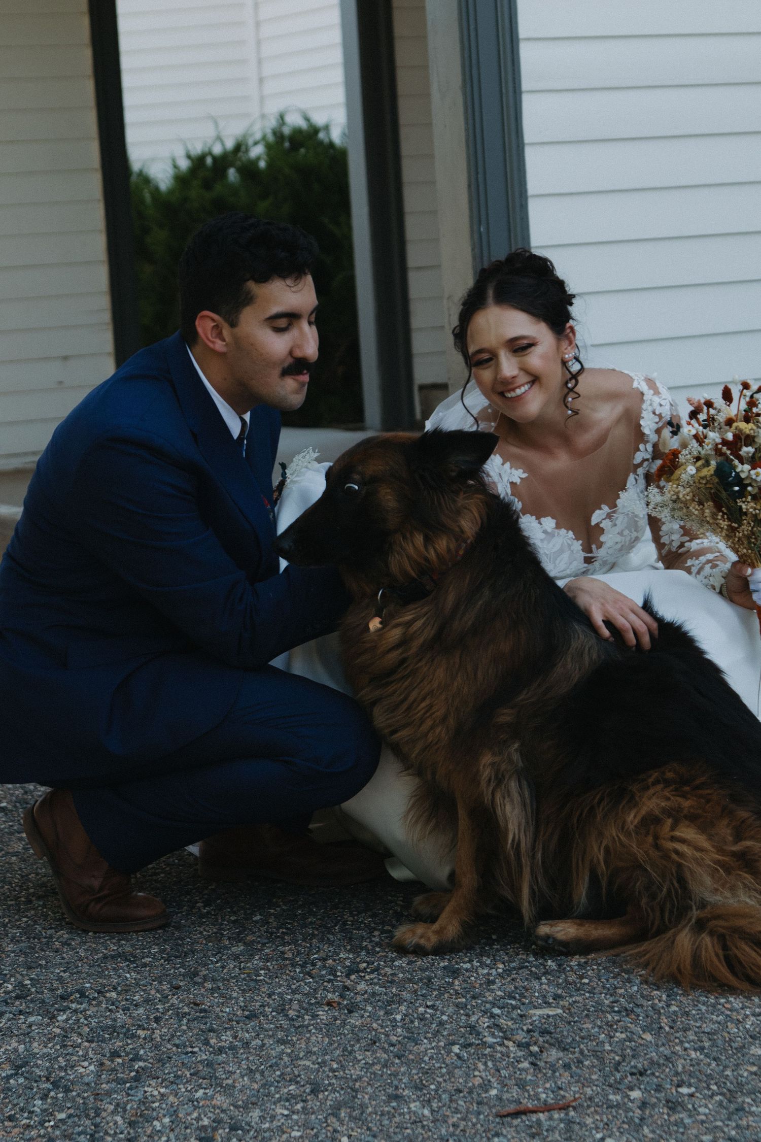 Newlyweds pose playfully with their dog on gravel ground during wedding photoshoot.
