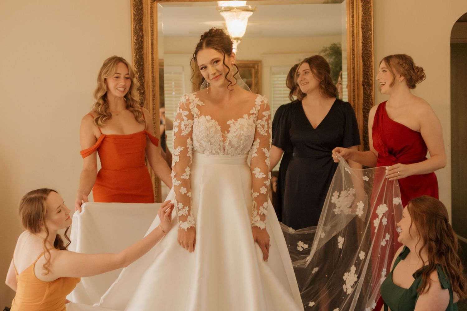 A bride in a lace-sleeved wedding gown surrounded by attendants in red and dark dresses.