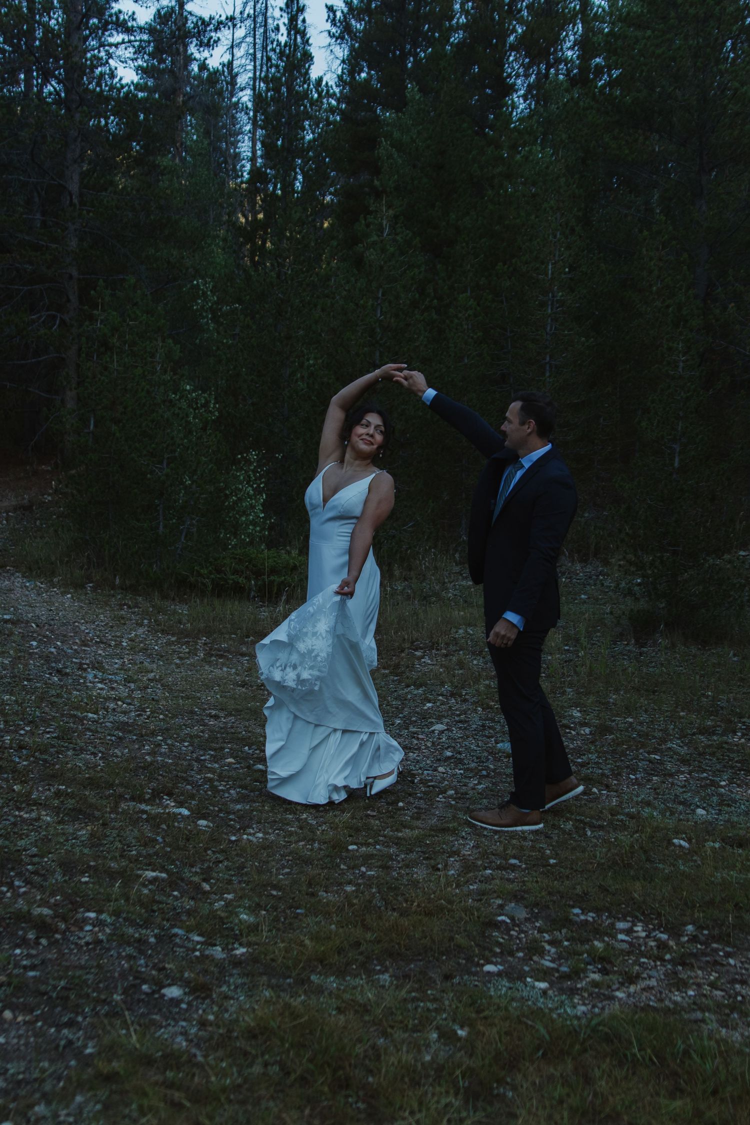 A wedding couple playfully dances in a forest clearing at dusk.