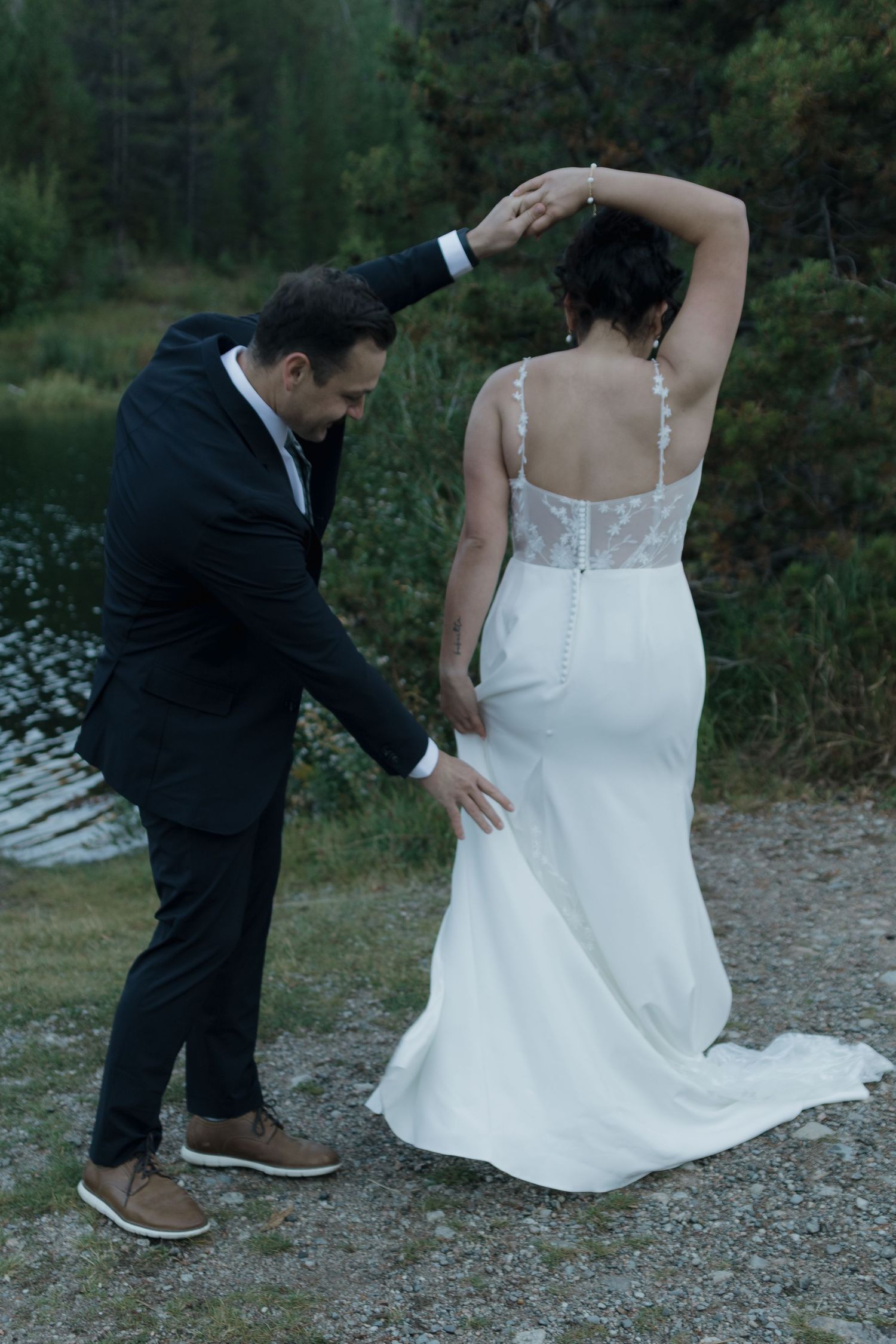 Groom helping adjust bride's white wedding dress on outdoor rocky terrain.