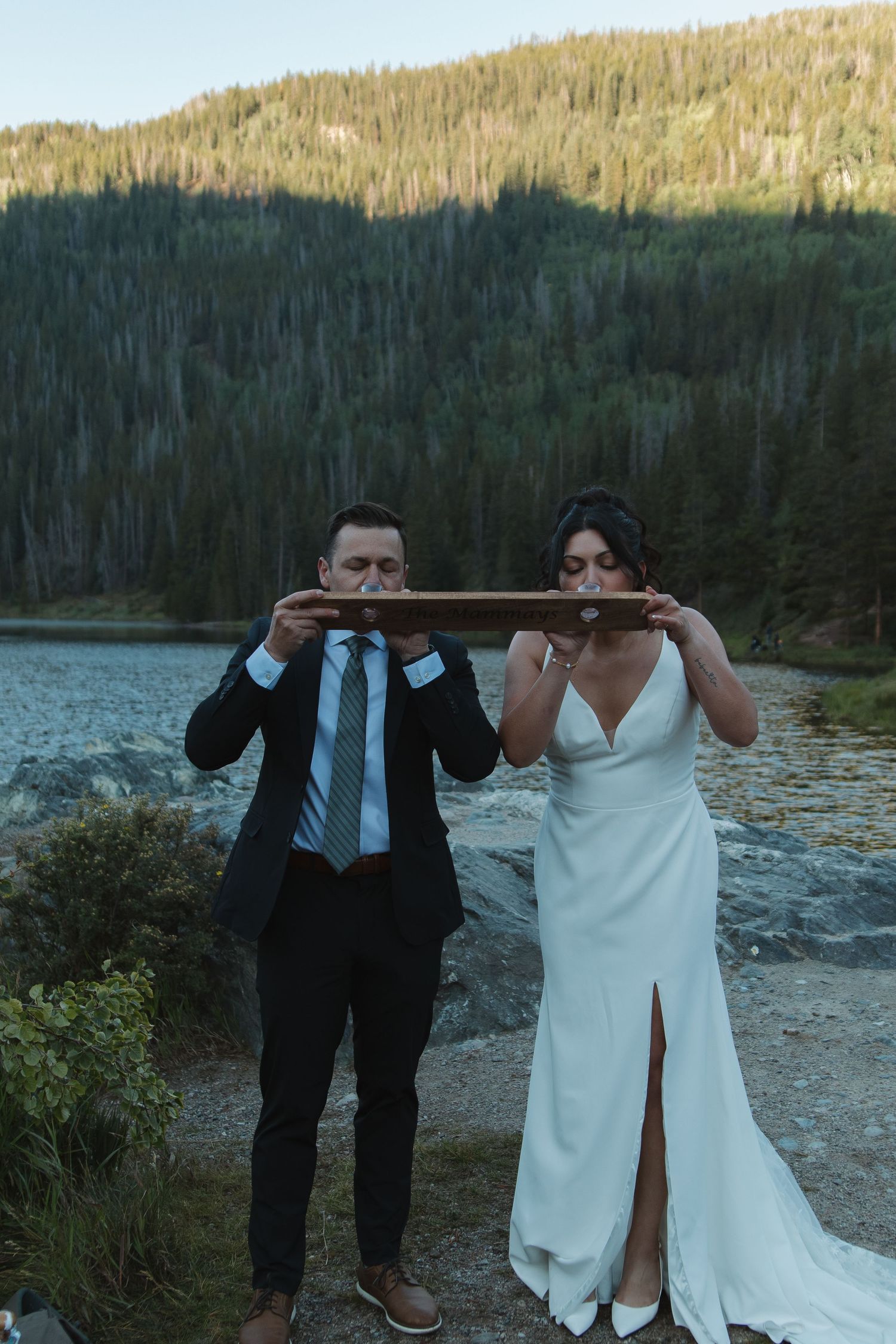 A joyful wedding couple shares a light-hearted moment on rocky terrain near a mountain lake.