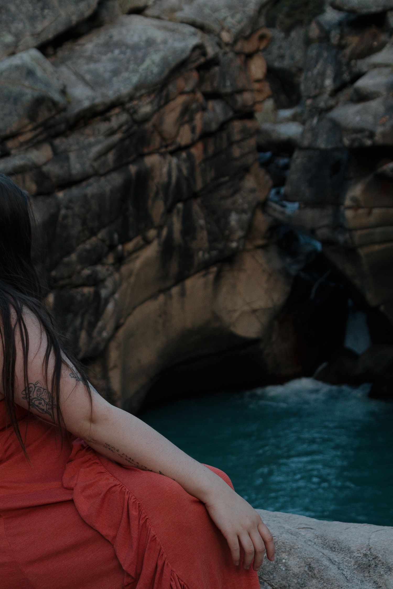 A person in a red dress sits near turquoise waters inside a dimly lit cave with dramatic rock formations.
