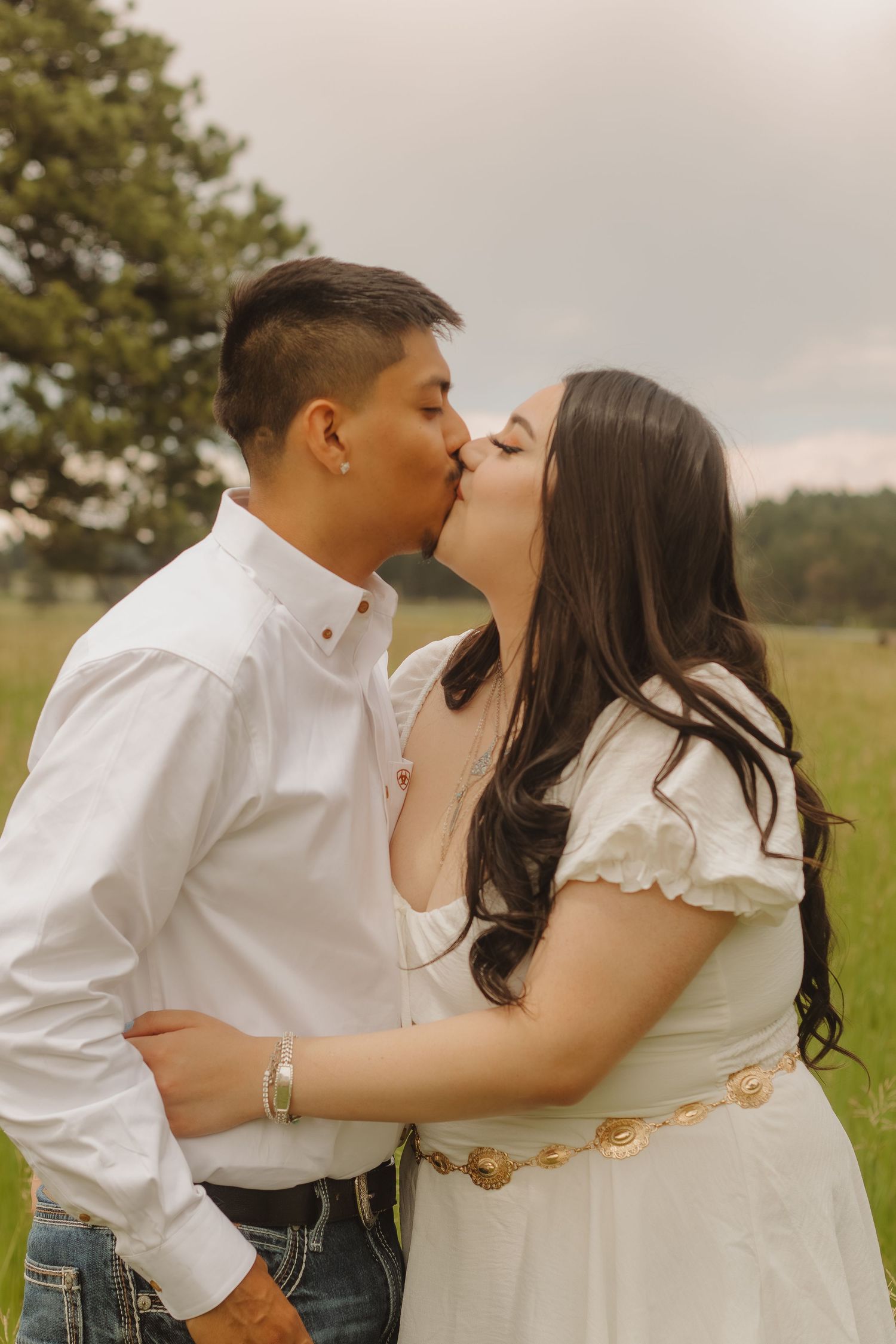 Couple sharing a romantic kiss in a grassy field with mountains in the background.