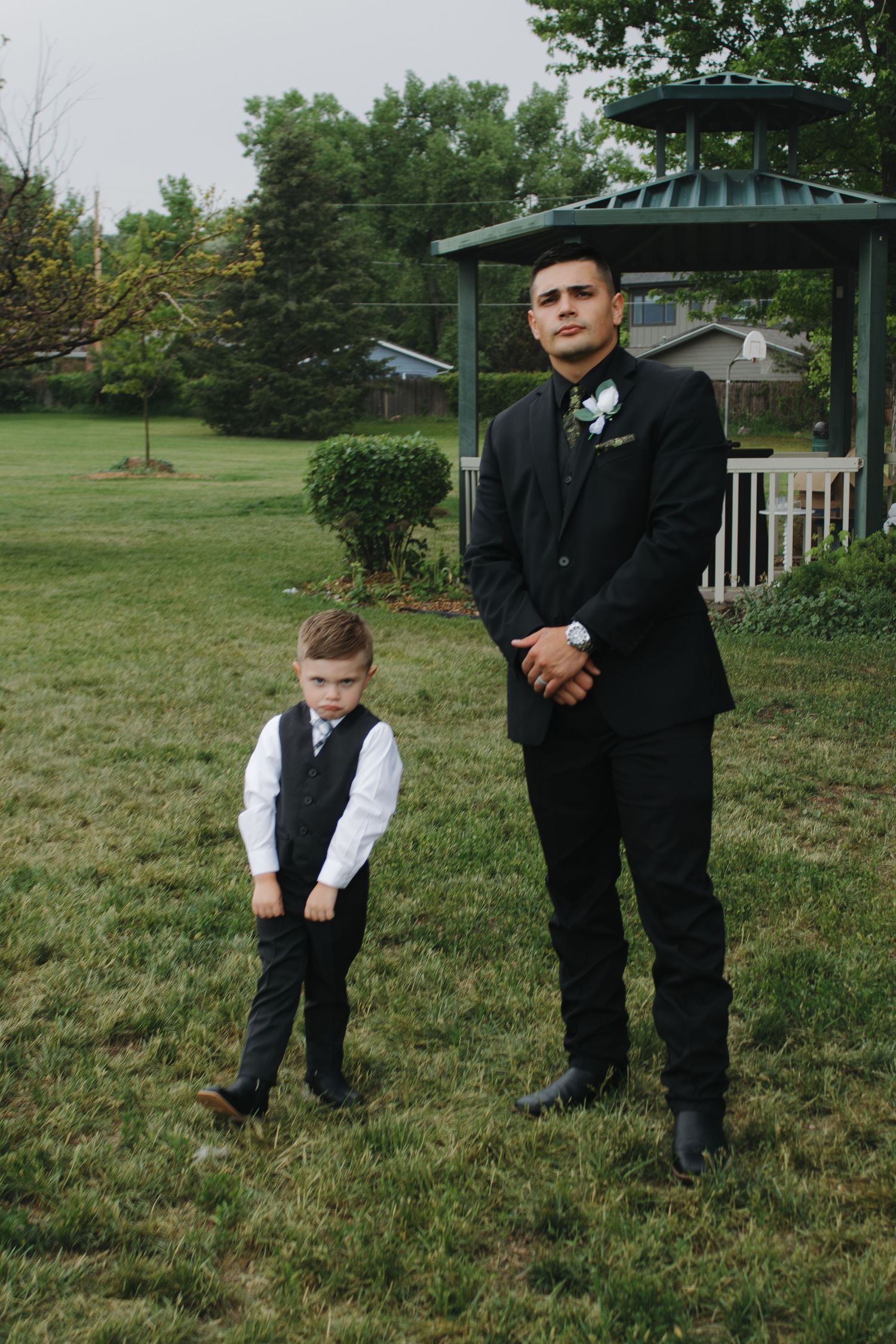 Young ring bearer in formal attire plays on grass near gazebo.