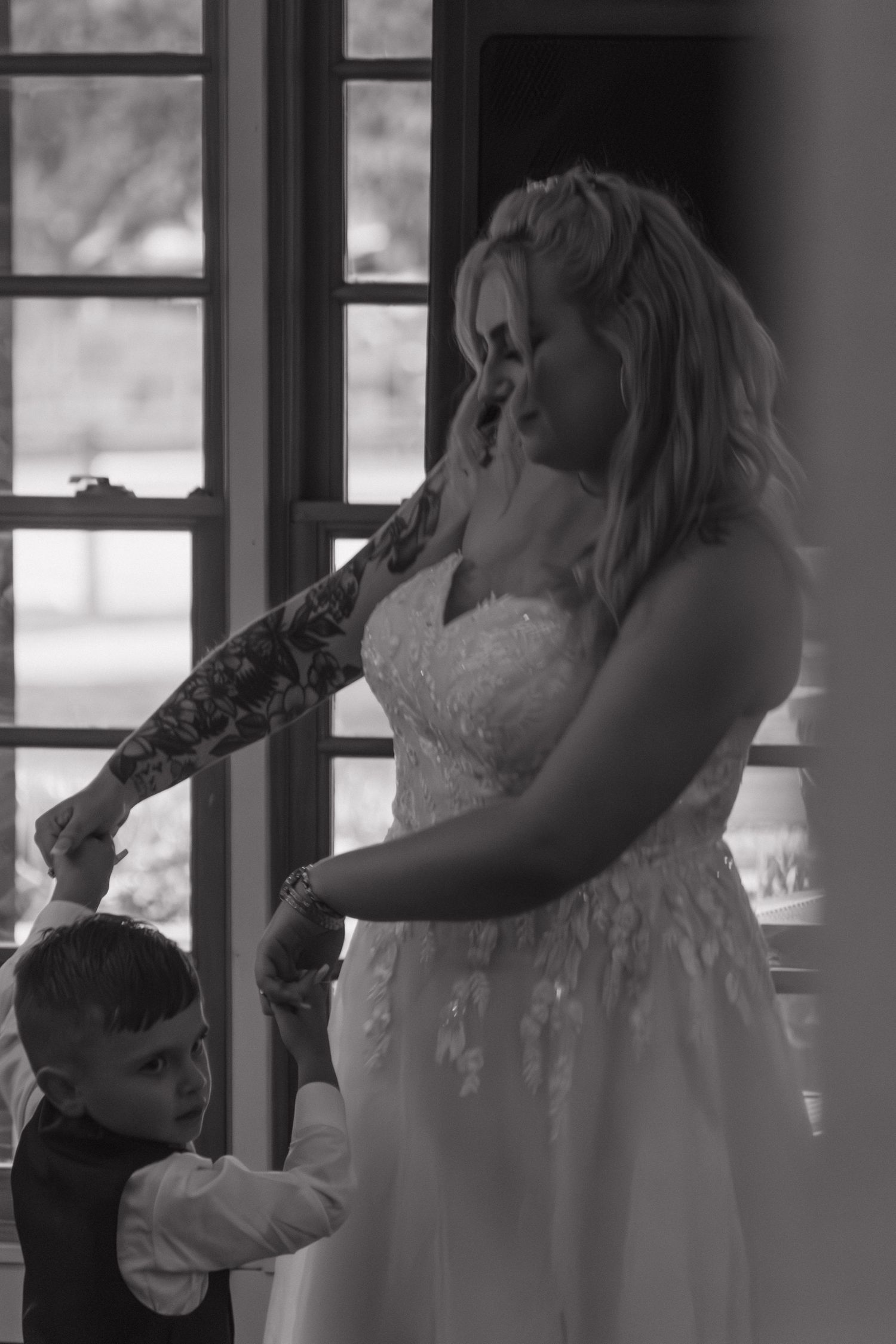 A bride in a white lace dress dances with a young boy in formal wear by a window.