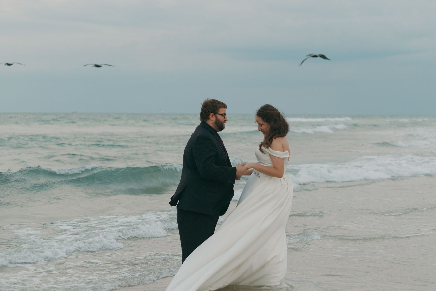 Couple walking along a beach with waves and seabirds in the background.