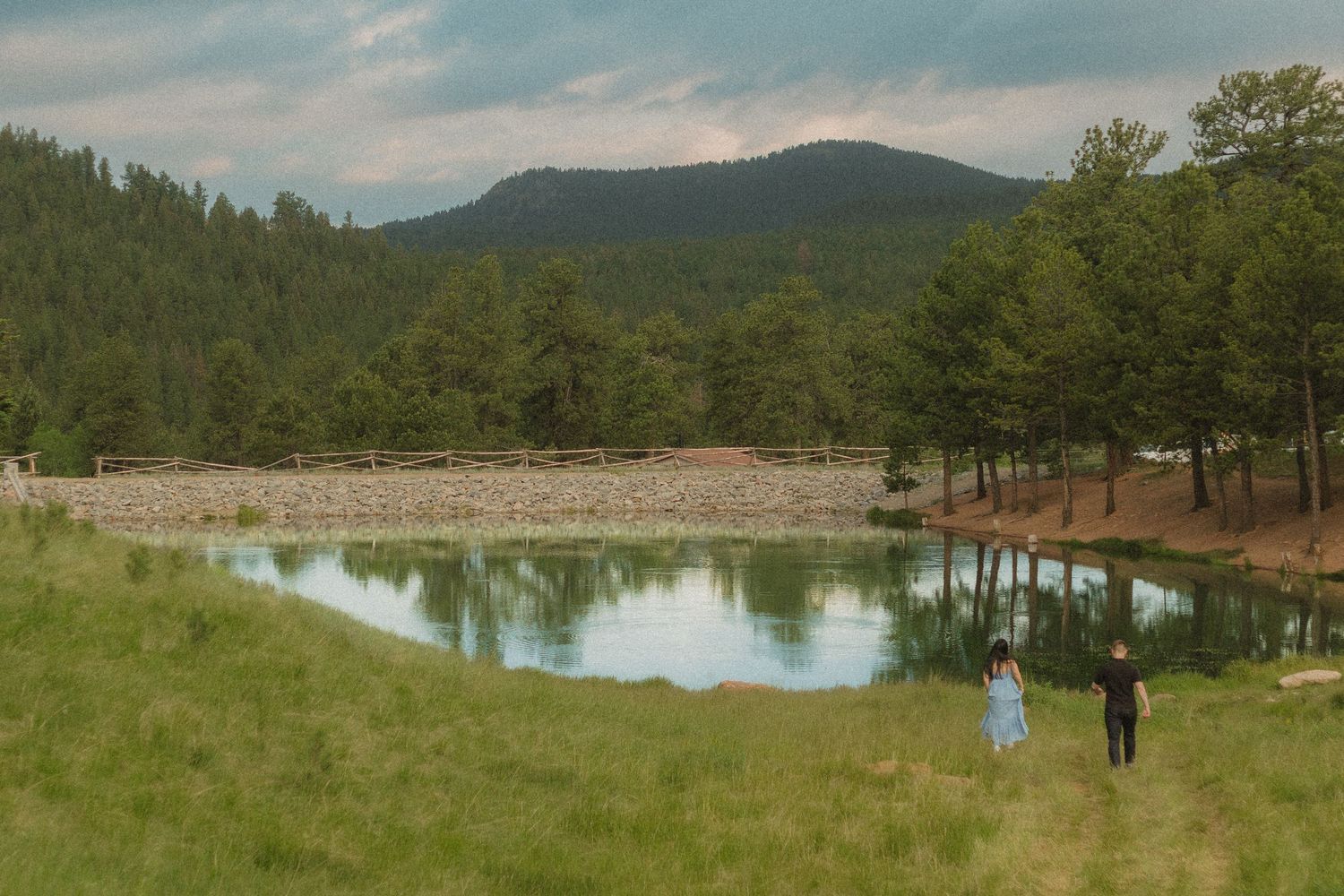 A couple stands near a mountain lake surrounded by pine trees under a cloudy sky.