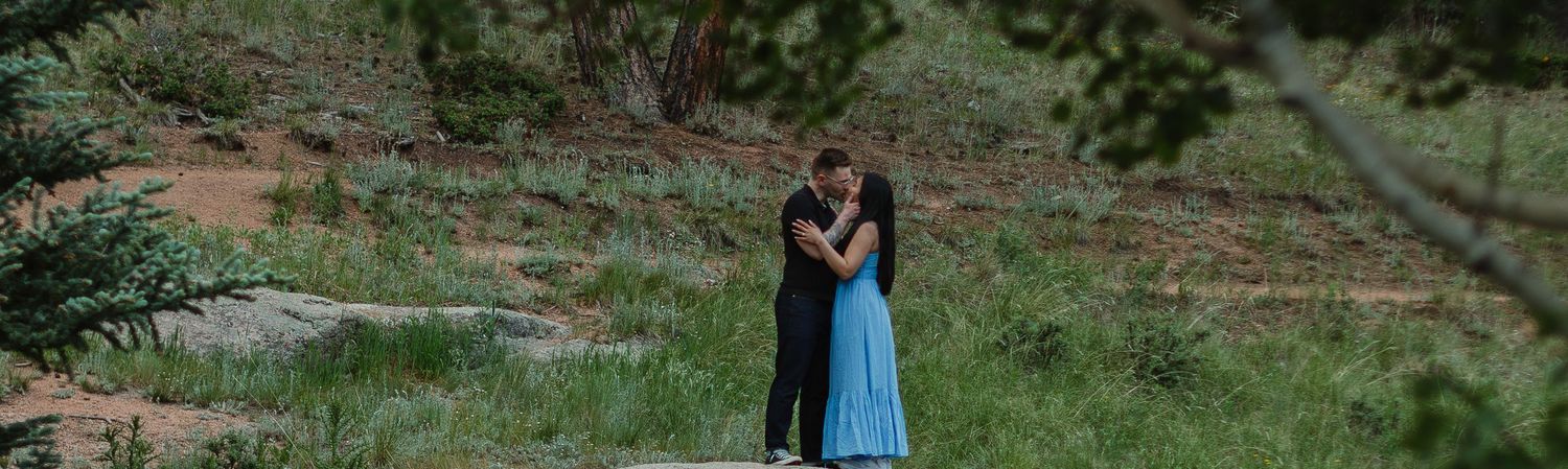 A romantic couple standing on a large boulder against a backdrop of evergreen forest.