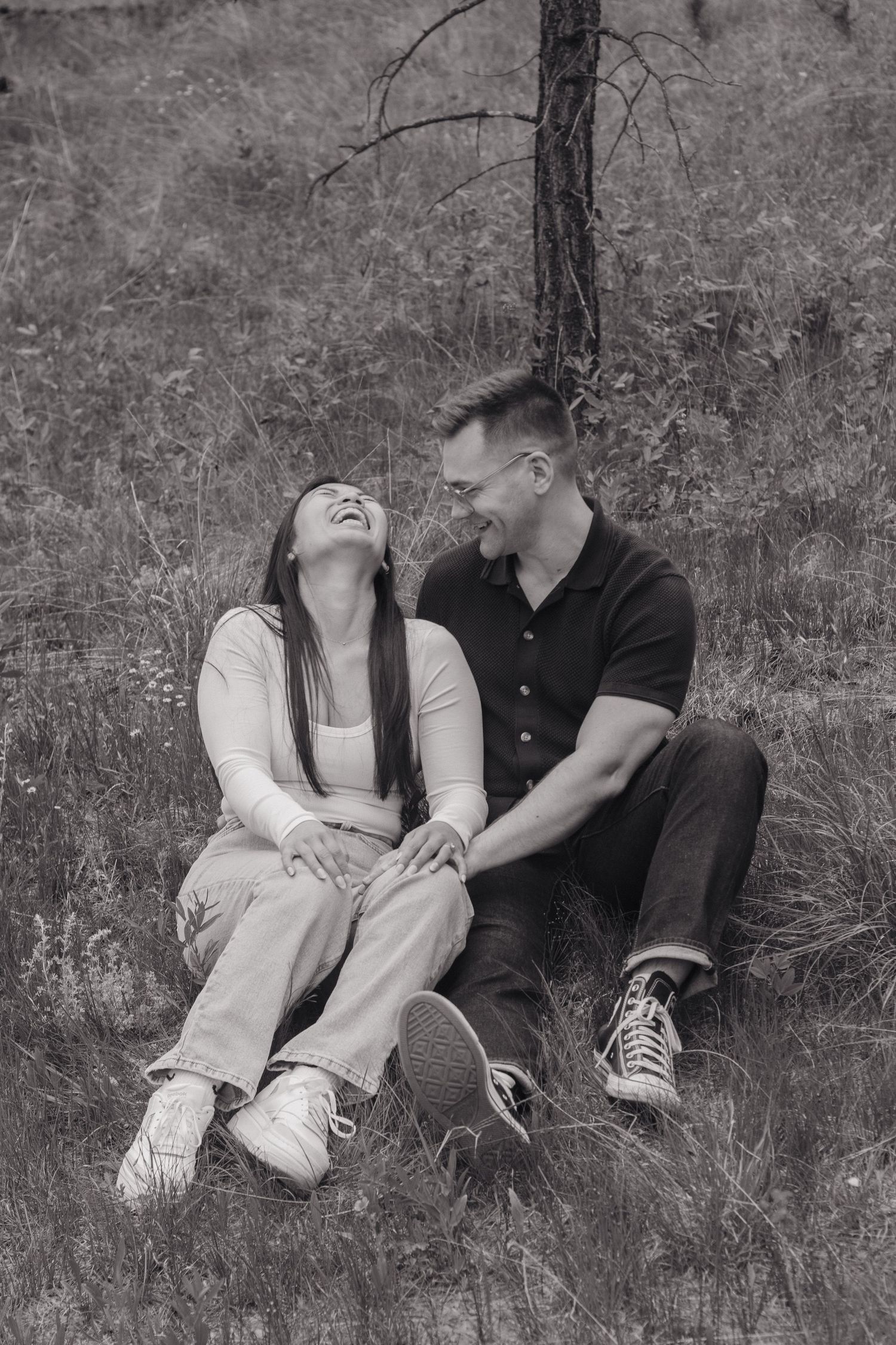 A black and white photo of a couple laughing together while sitting on a grassy hillside.