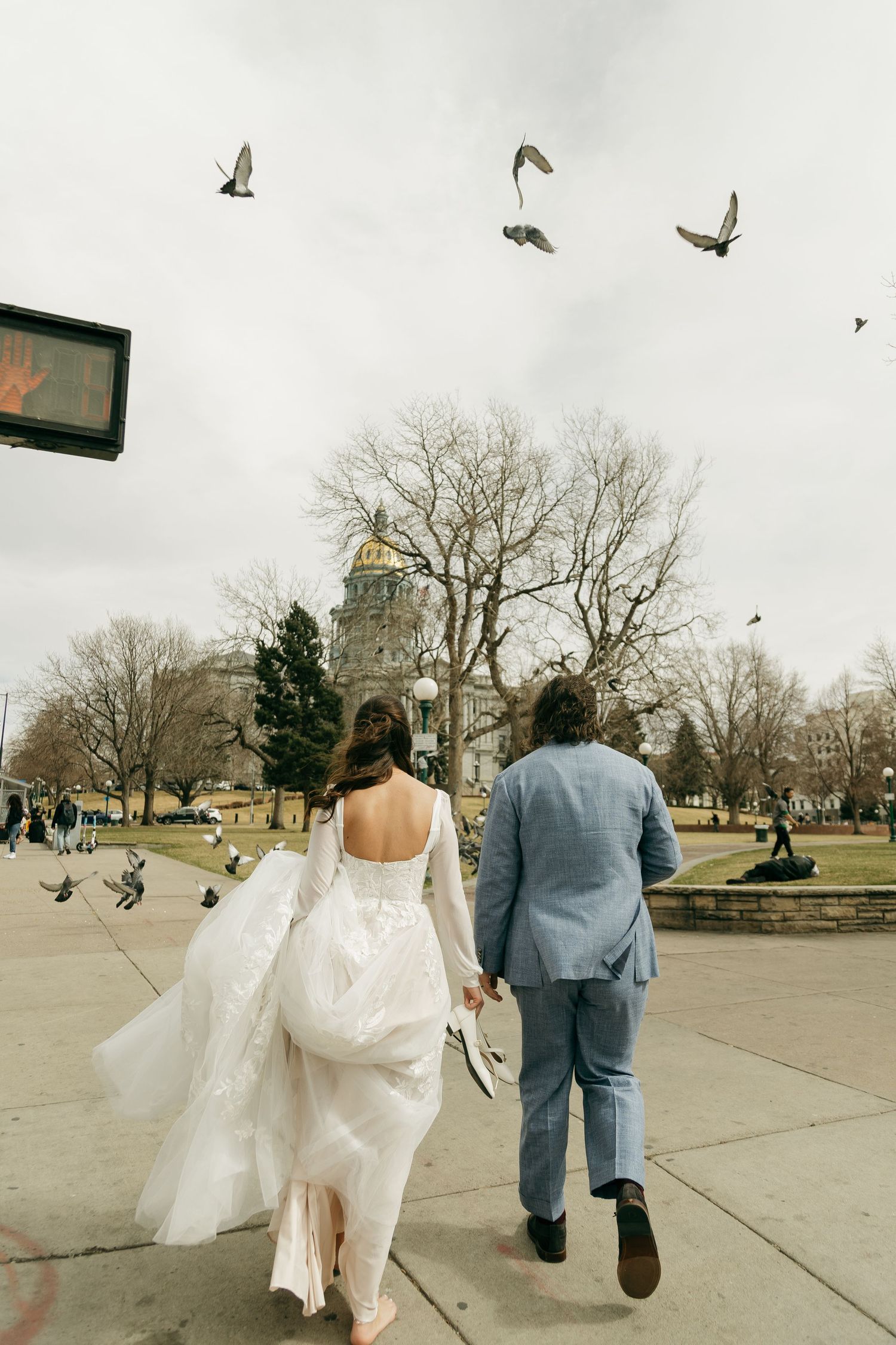 A couple in wedding attire walks across a plaza with flying pigeons and a church spire in the background.