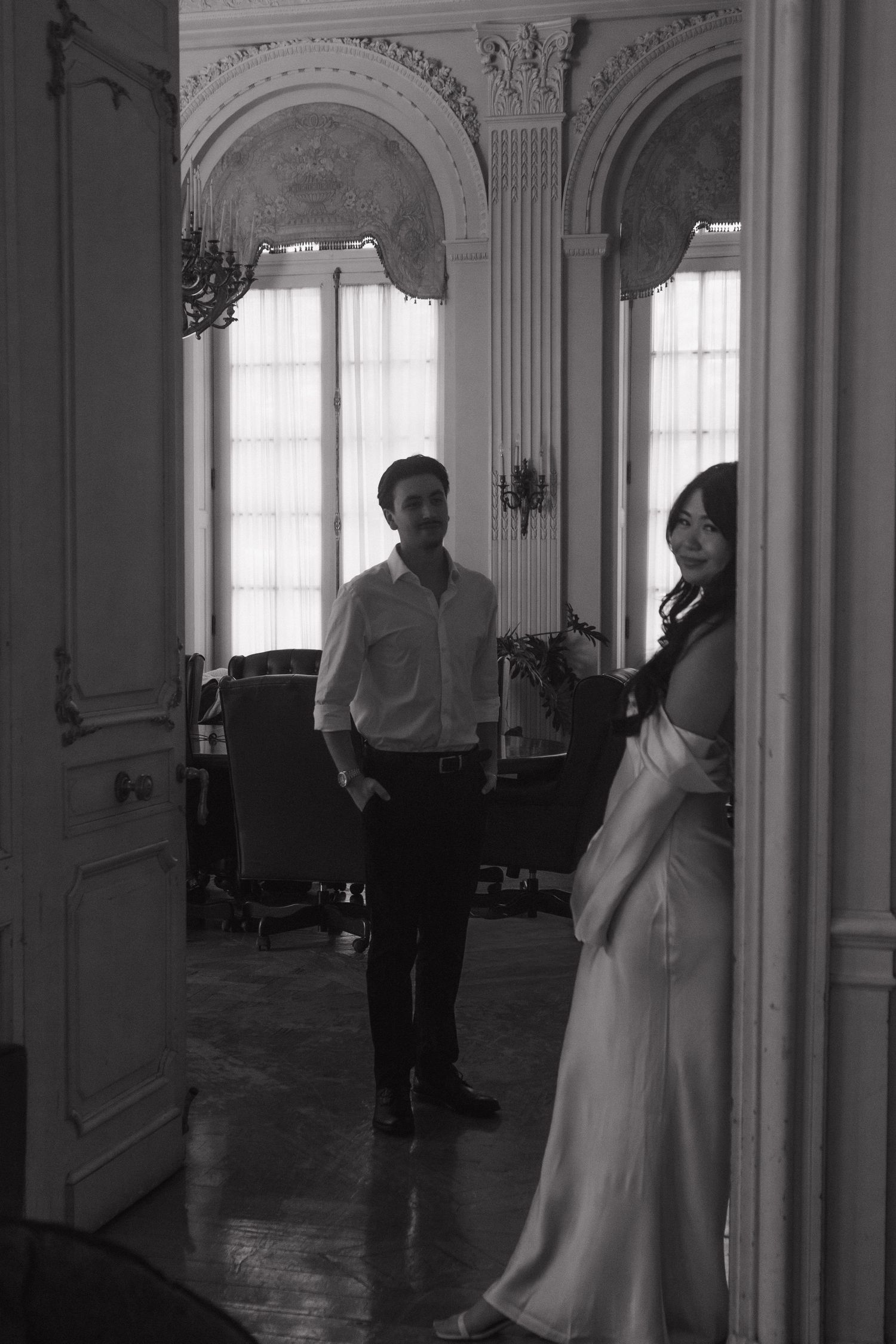 A couple in formal attire stands in an elegant white hallway with ornate architectural details.