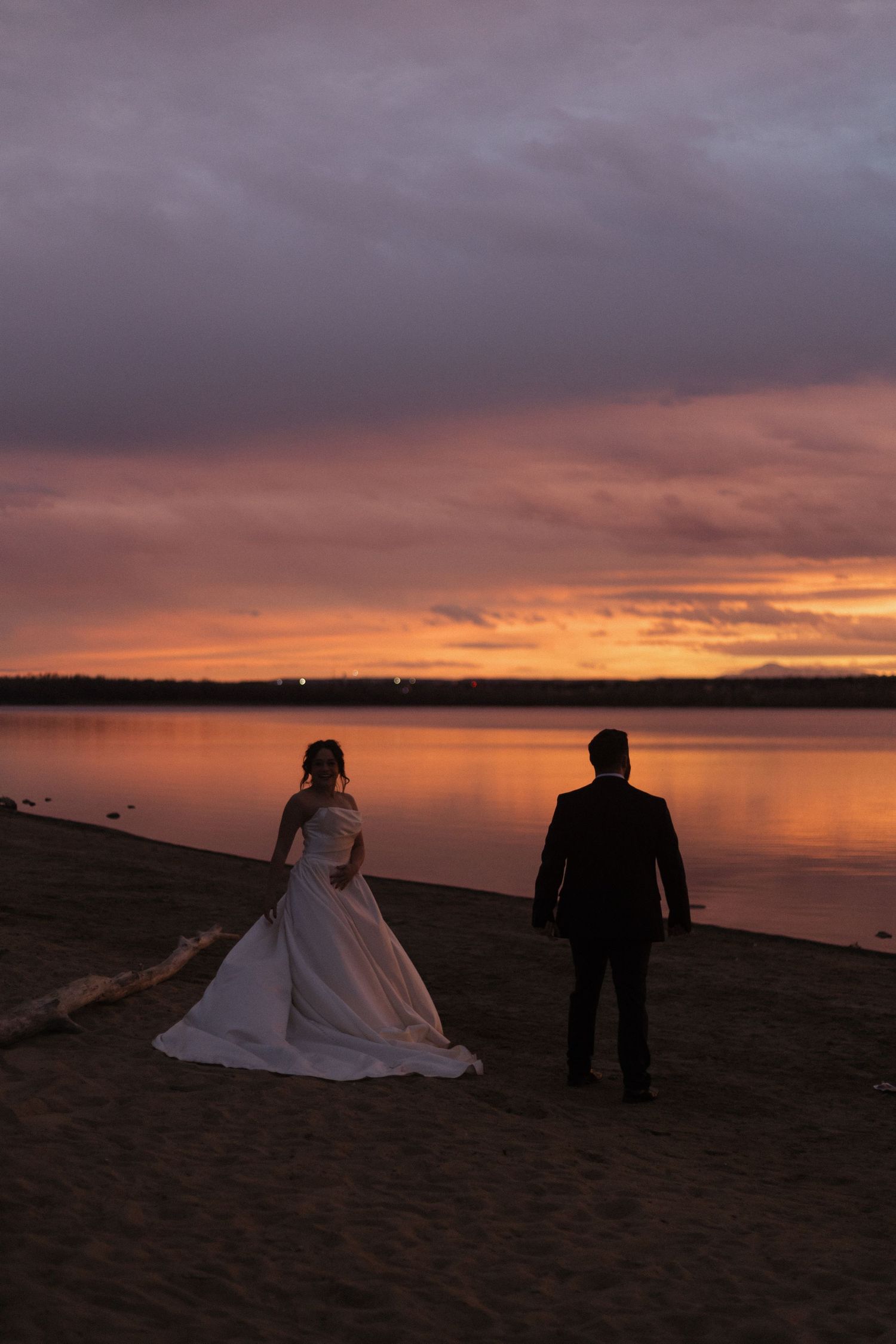 Silhouettes of a couple in formal attire stand on a beach at sunset with dramatic orange and purple clouds.