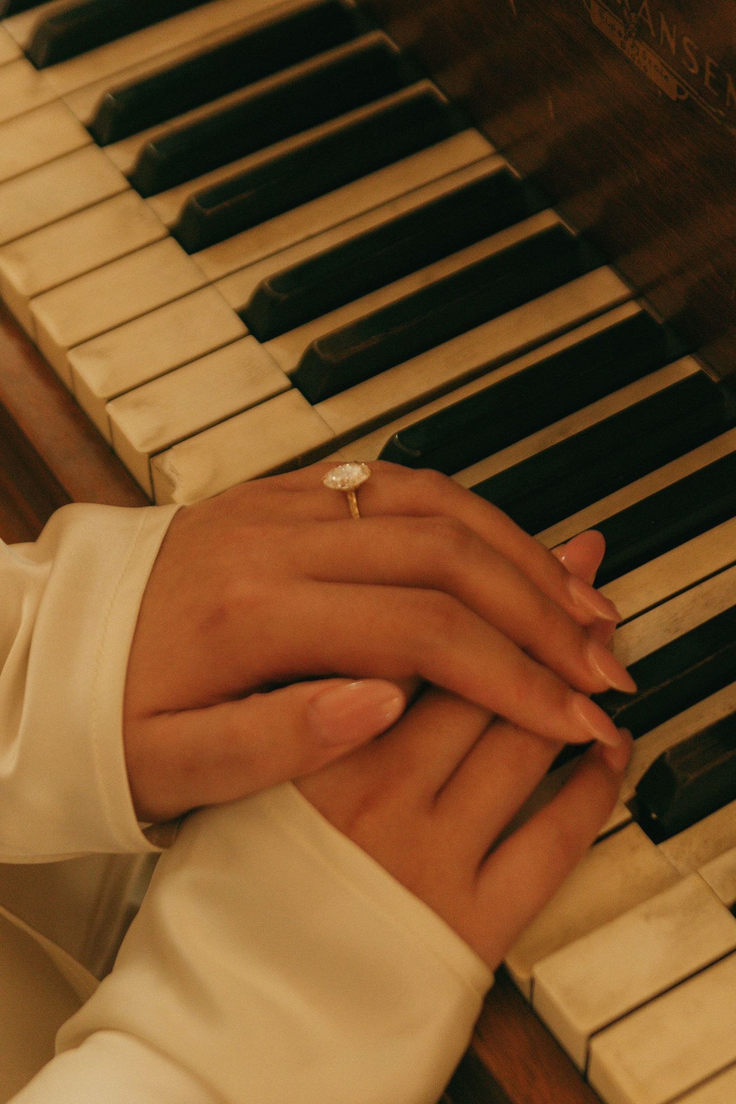 Hands wearing white sleeves rest gently on piano keys with an engagement ring visible.
