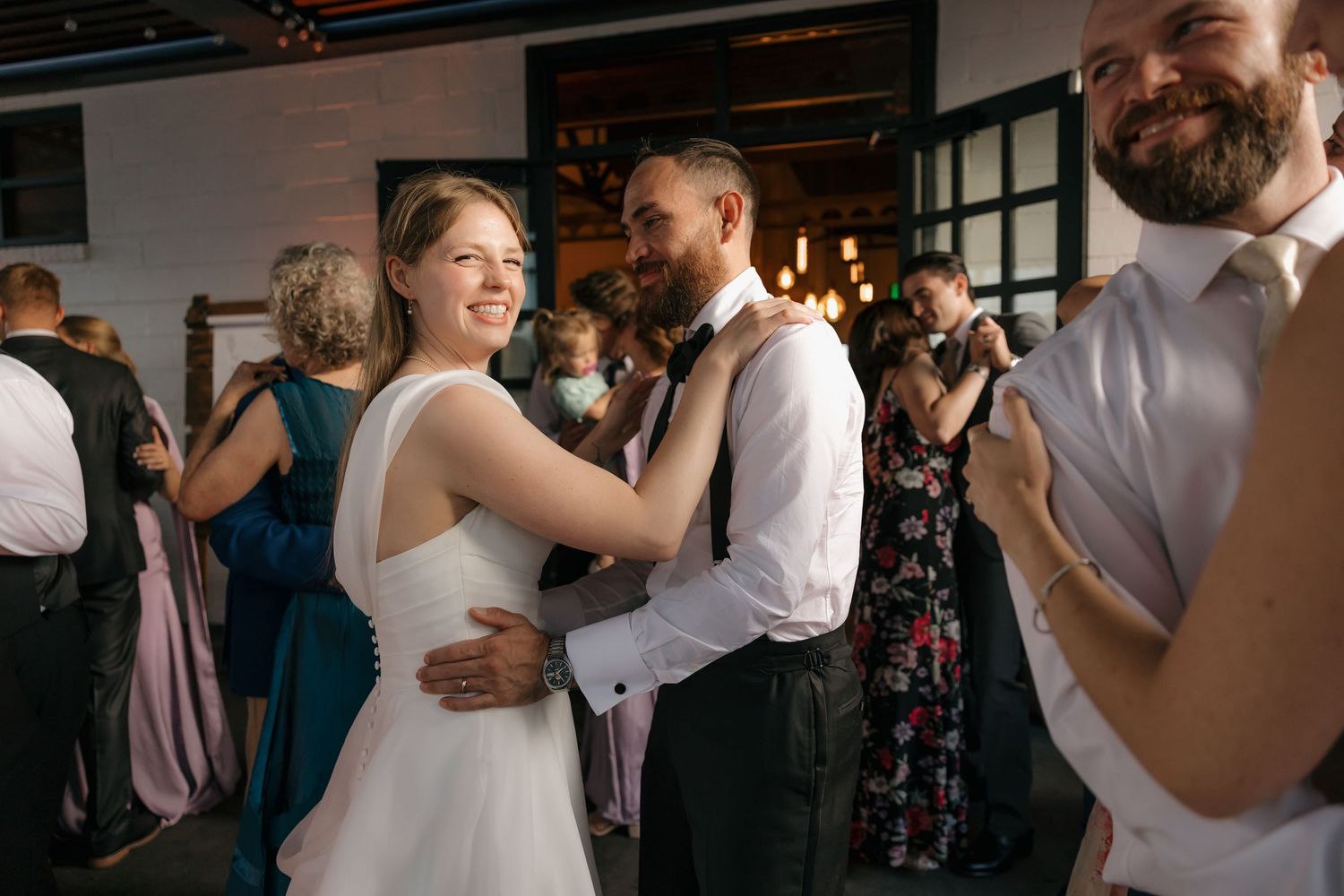 Bride in backless white gown shares first dance with groom in suspenders at wedding reception.
