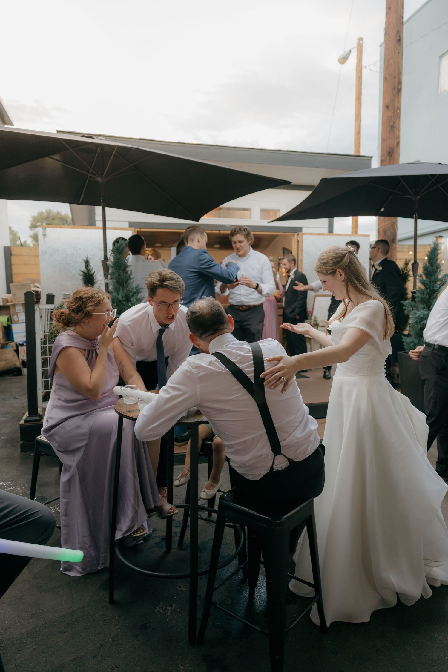 Bride in white gown mingles with guests at outdoor wedding reception under black patio umbrellas at dusk.