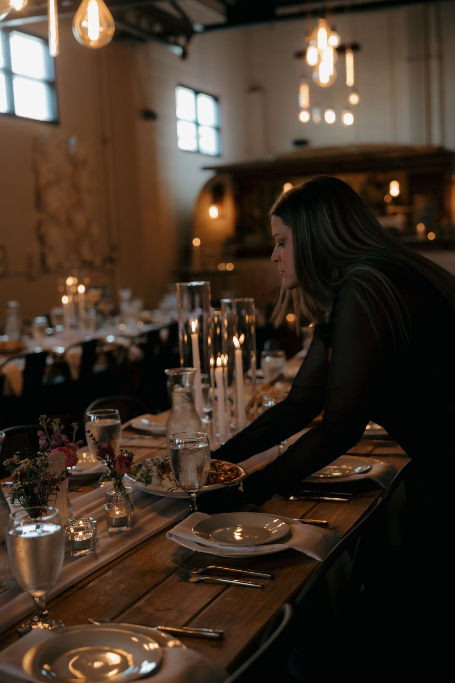 Wedding coordinator arranging elegant candlelit reception table in rustic industrial venue.