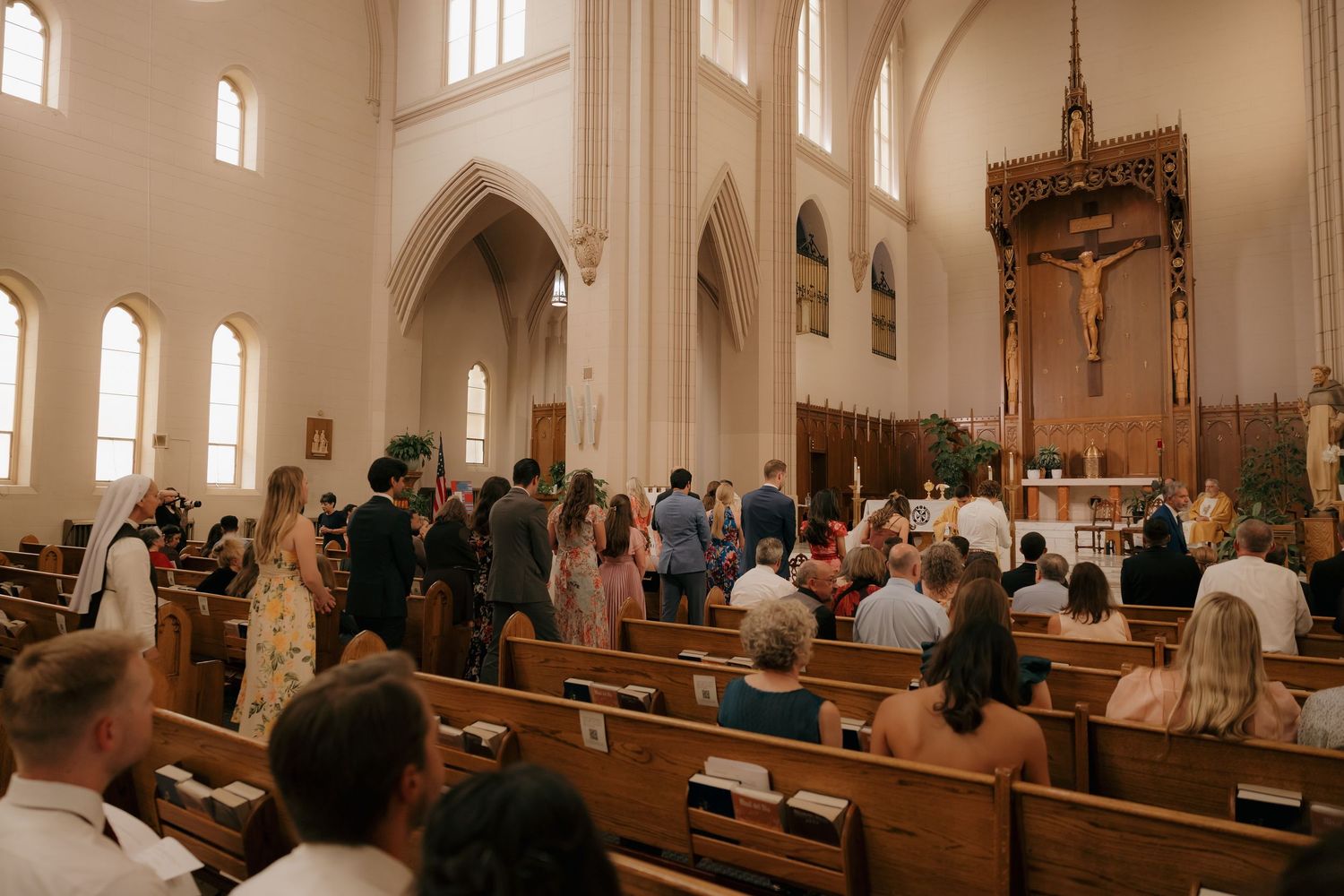 Guests standing in wooden pews during a Catholic church wedding ceremony with Gothic arches and altar crucifix.