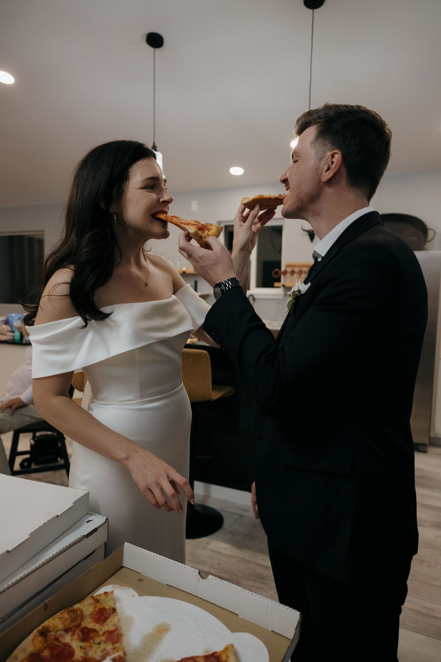 Bride and groom feeding each other pizza slices in wedding attire at their casual reception.
