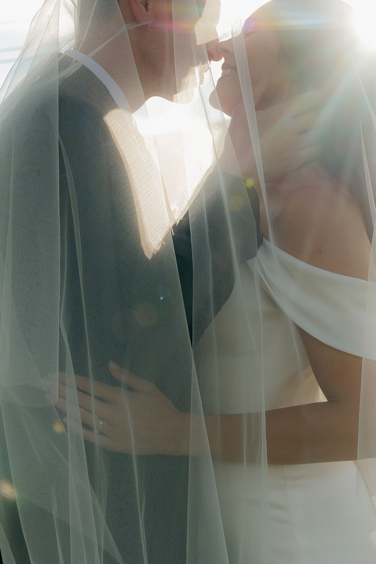 A bride and groom share an intimate moment under a flowing veil with soft sunlight creating a romantic lens flare.