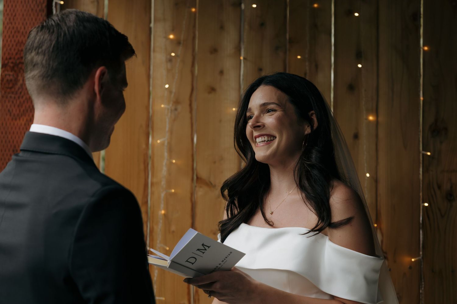 Bride in white off-shoulder dress smiling while reading vows, groom in gray suit, rustic wood backdrop with fairy lights.