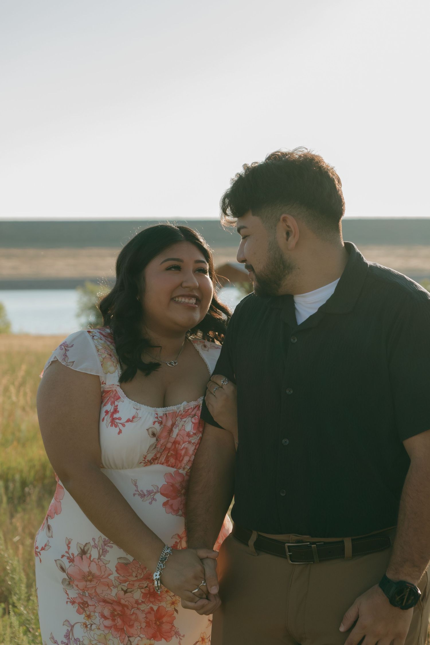 A couple shares an intimate moment at sunset in a field near a lake, with her wearing a floral dress and him in dark attire.