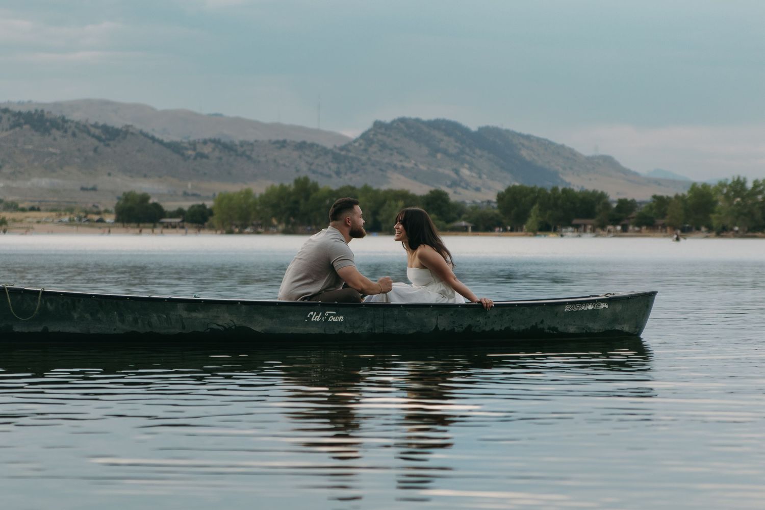 Two people share an intimate moment in a canoe on a tranquil lake surrounded by mountains.