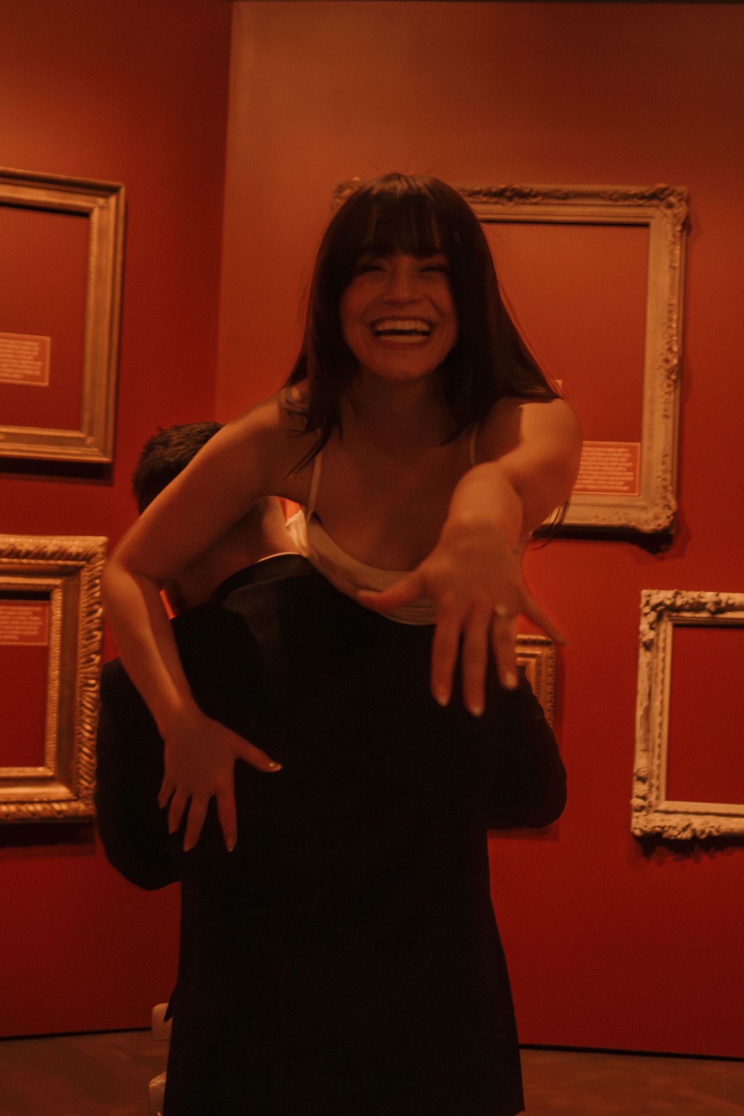 Figure in black dress poses playfully in front of red gallery wall with framed artwork.