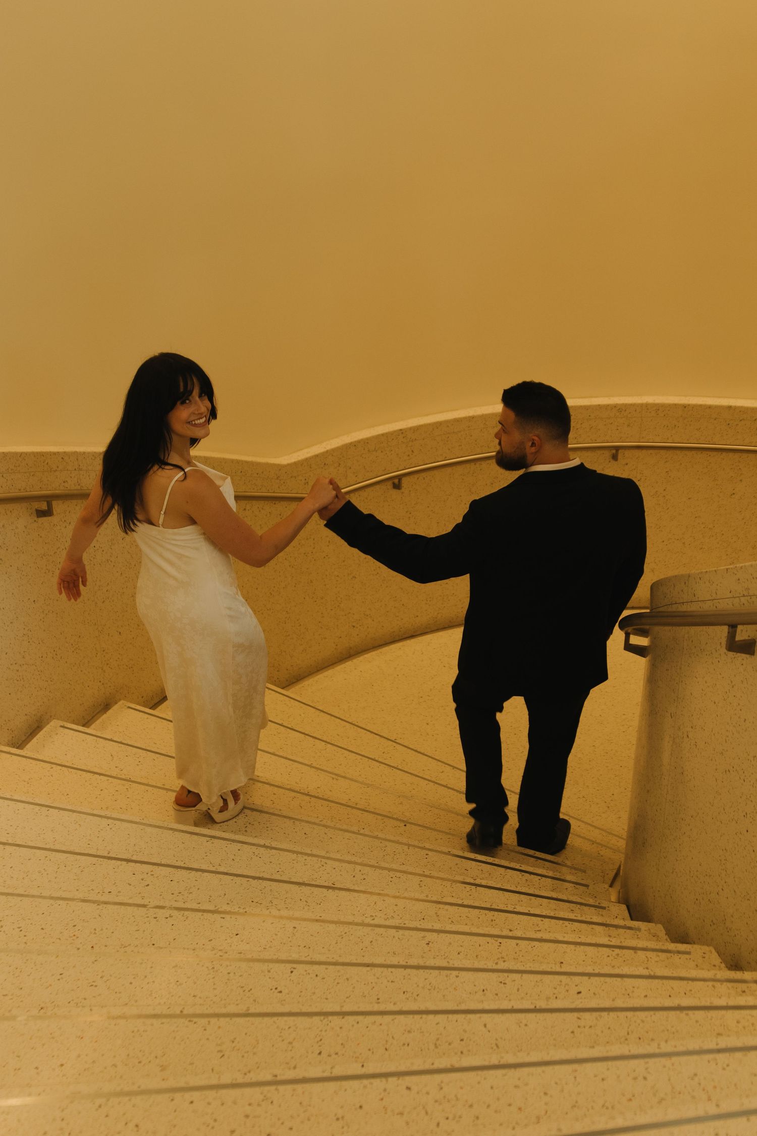 A couple descends a curved marble staircase in elegant formal attire.