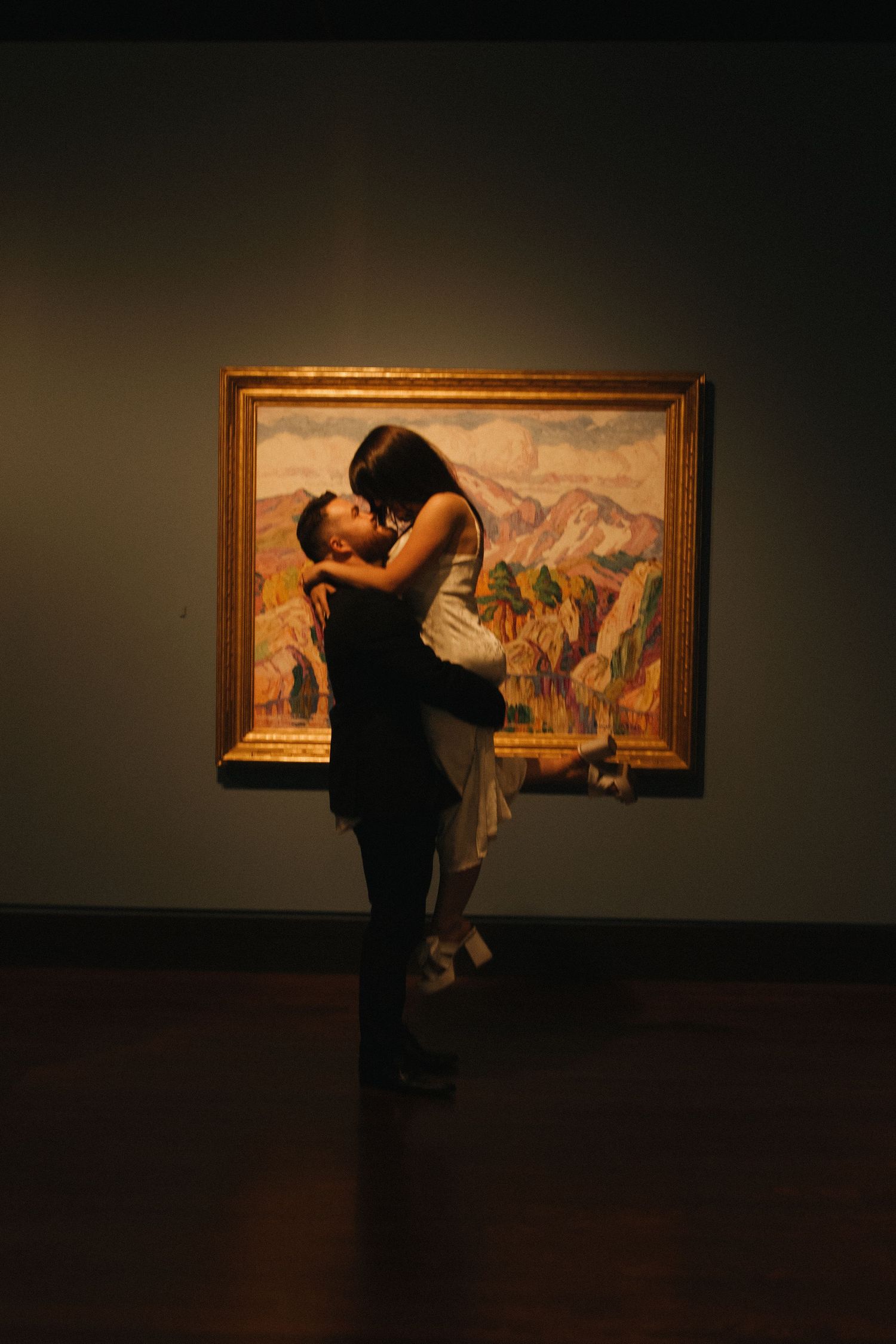 Silhouetted figures embrace in front of illuminated artwork in a dark museum gallery.
