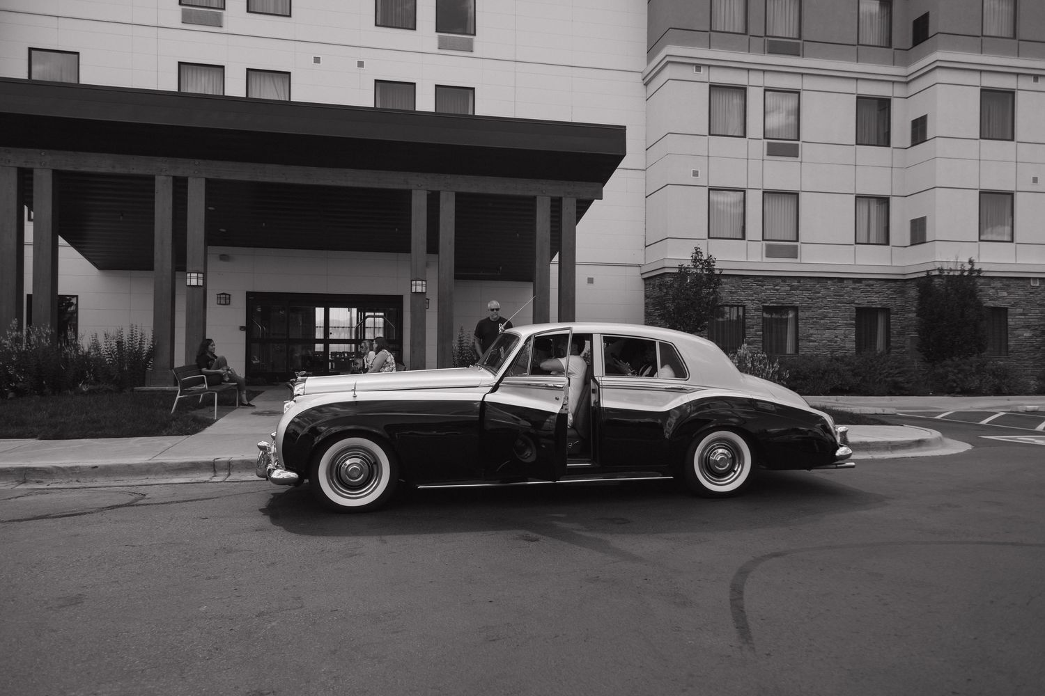 Vintage black and white classic car parked outside a hotel entrance in a black and white photograph.