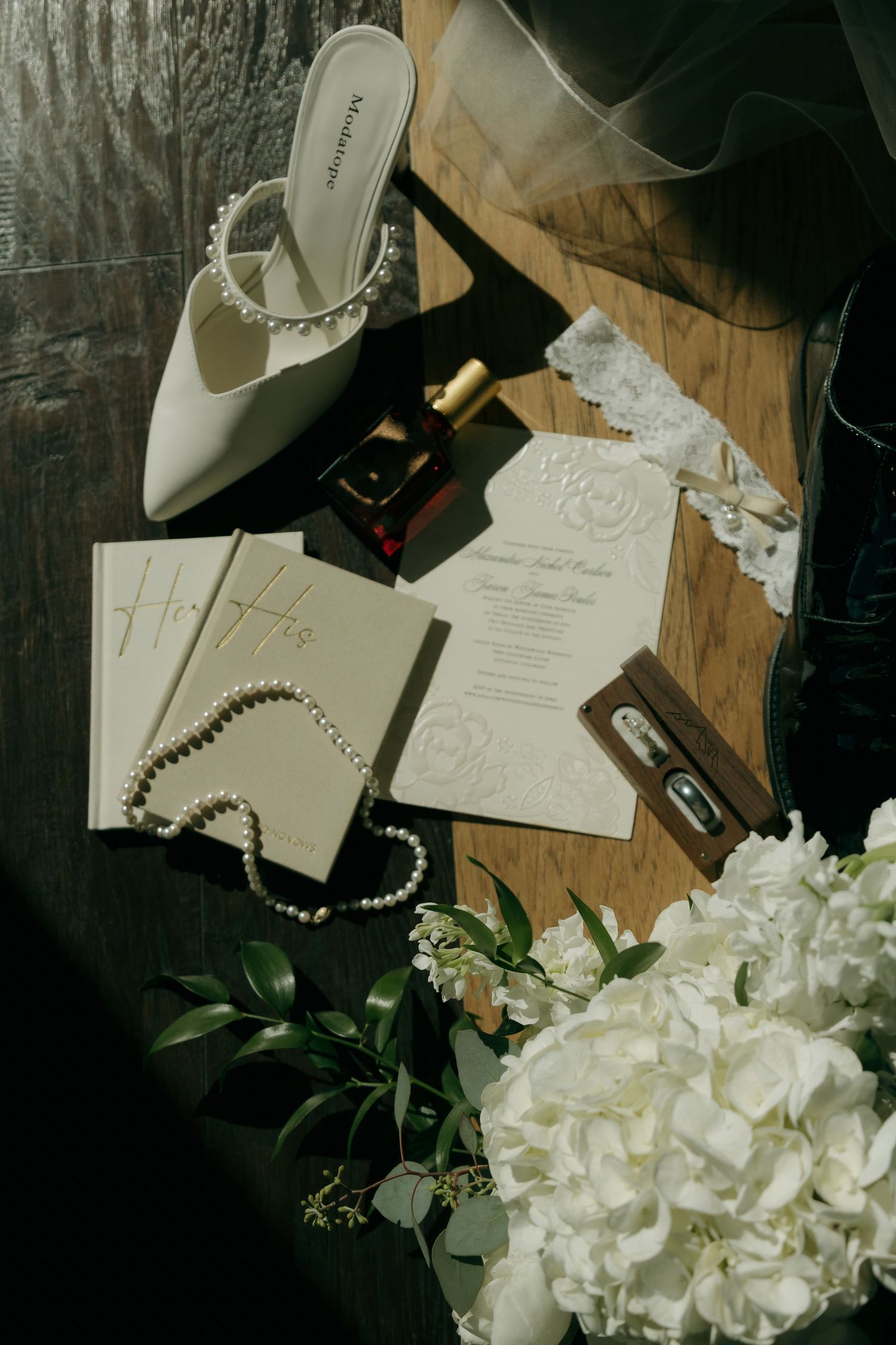 Flat lay of wedding details including pearl heels, bouquet, invitation, perfume, and jewelry.